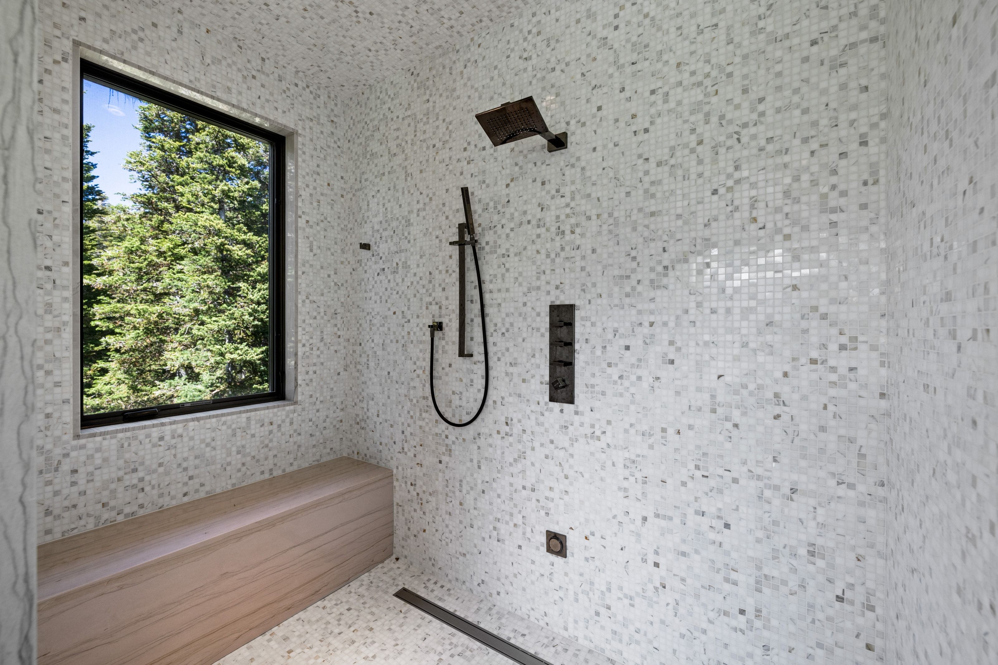 Shower featuring mosaic tile walls and floor, a fixed window, a built-in wood-finish bench, and matte black fixtures including a rainfall showerhead and handheld sprayer