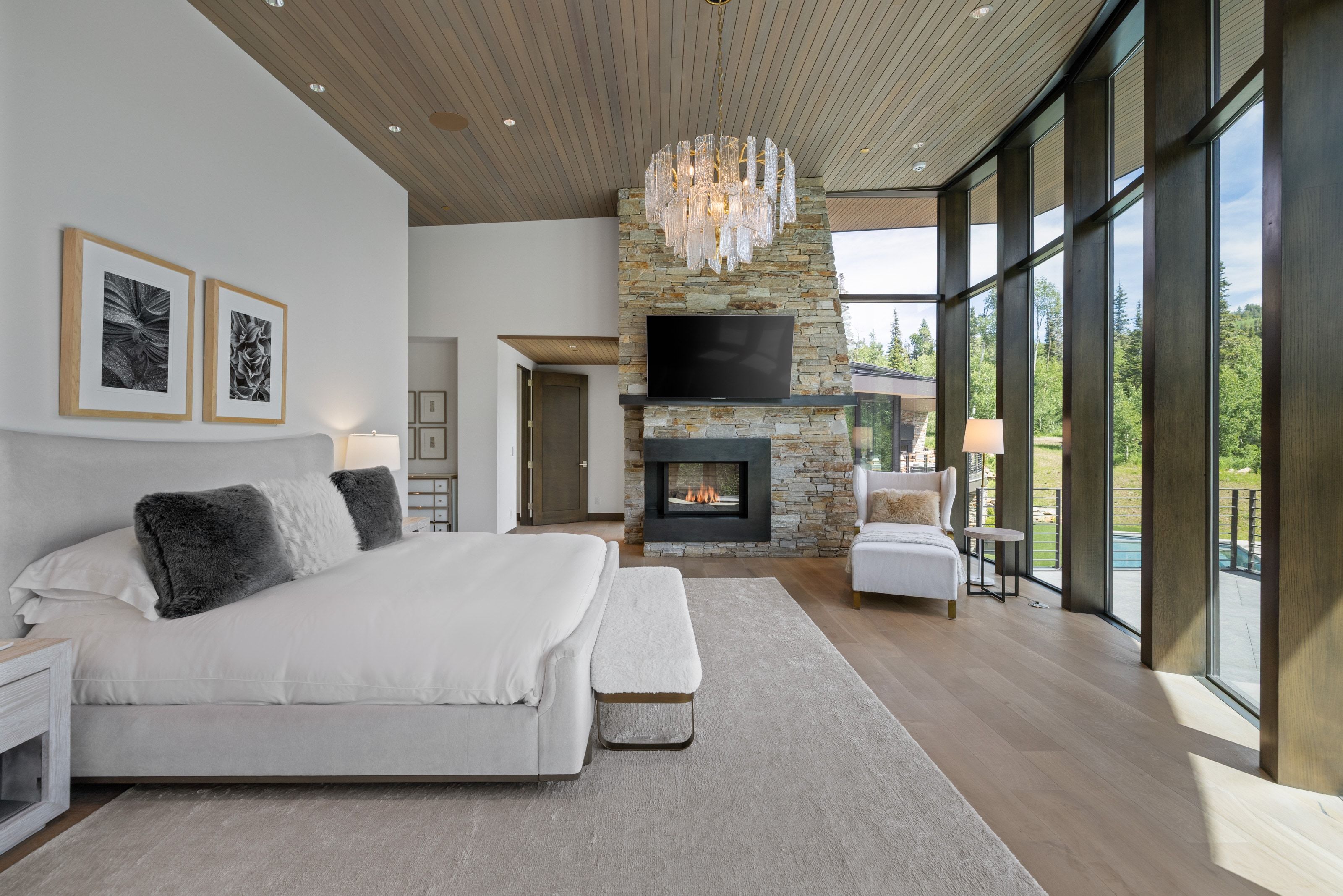 Floor-to-ceiling windows with dark wood-finish frames, a stacked stone fireplace with an integrated television, wood-finish flooring, a light wood-paneled ceiling, and a prominent decorative chandelier