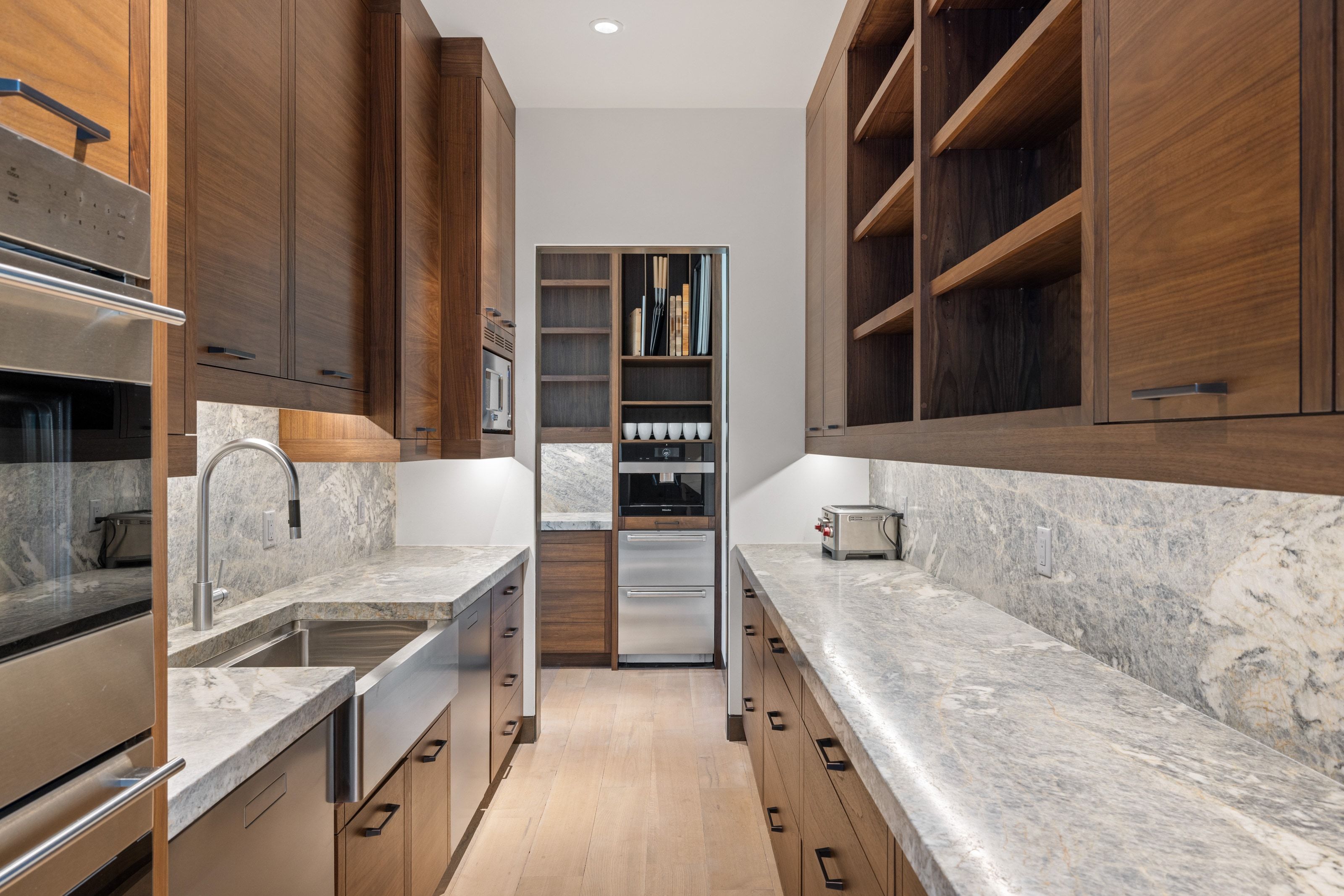 Galley kitchen featuring wood-finish cabinetry, stainless steel appliances, stone countertops, and a farmhouse sink