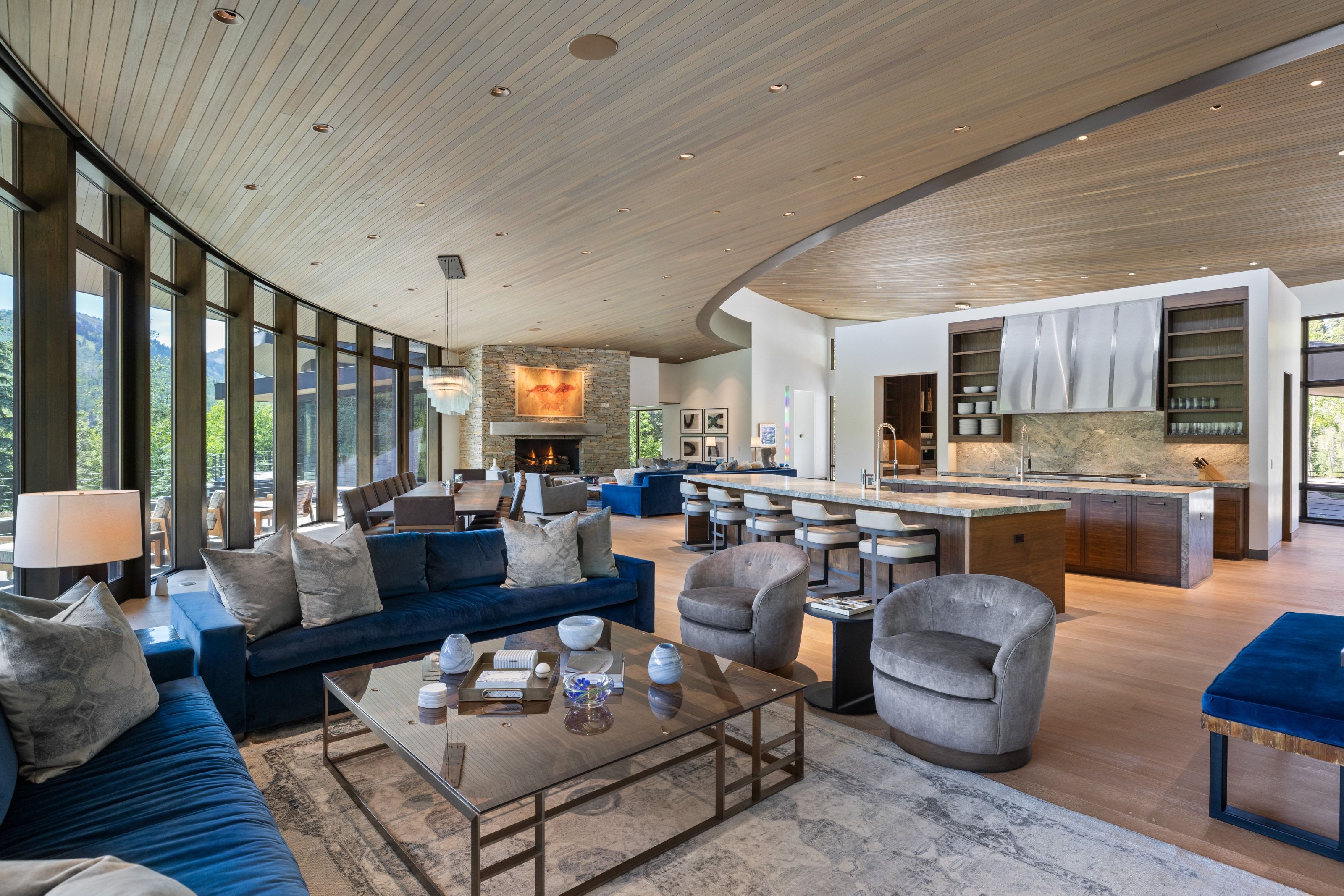Spacious open-concept living area featuring a curved wall of floor-to-ceiling windows, a stone-clad fireplace, and a wood-slat ceiling with integrated lighting