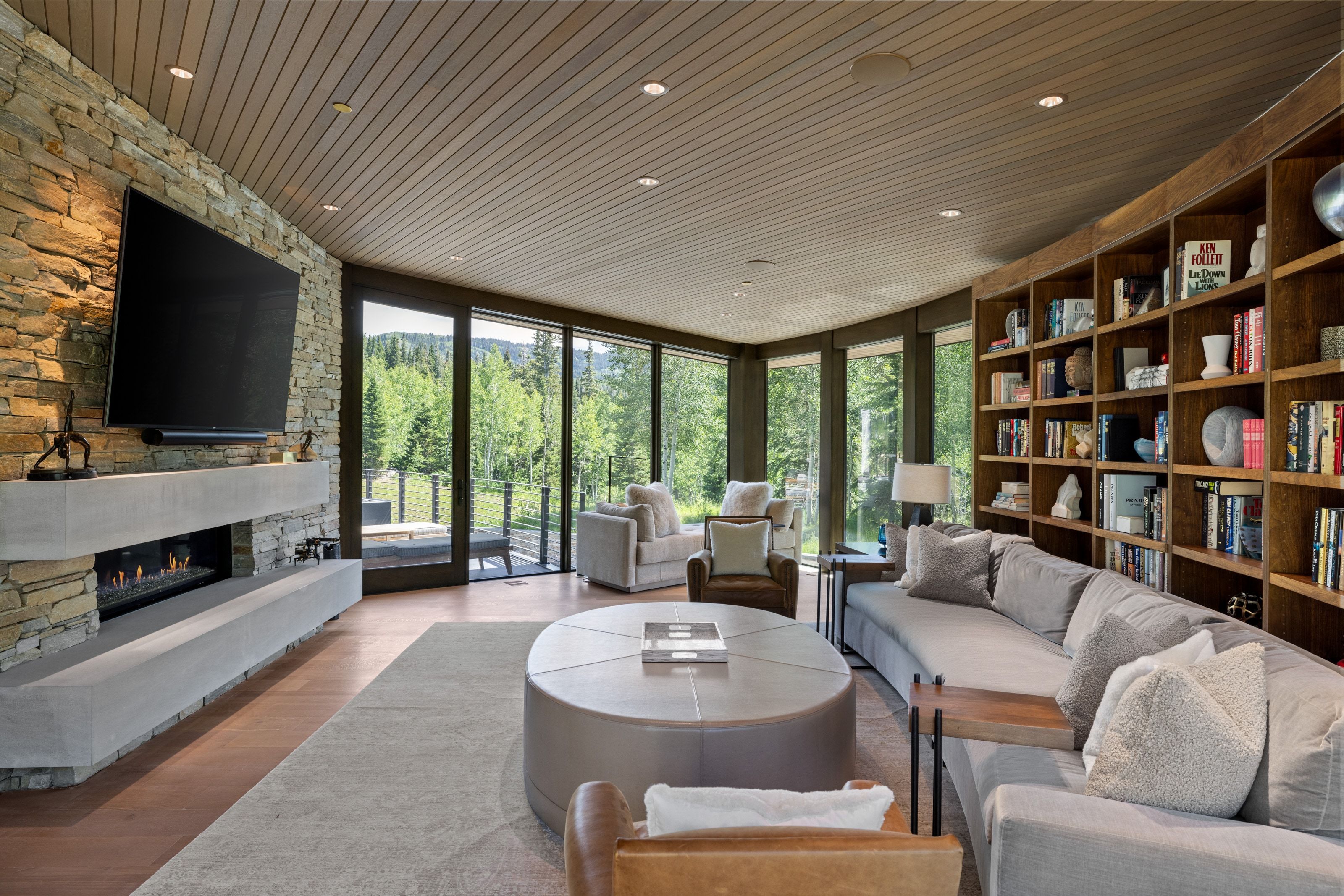 Spacious living area featuring a full-height stone accent wall with a linear fireplace, custom built-in shelving, wood-finish flooring, and large glass panels overlooking a treed landscape