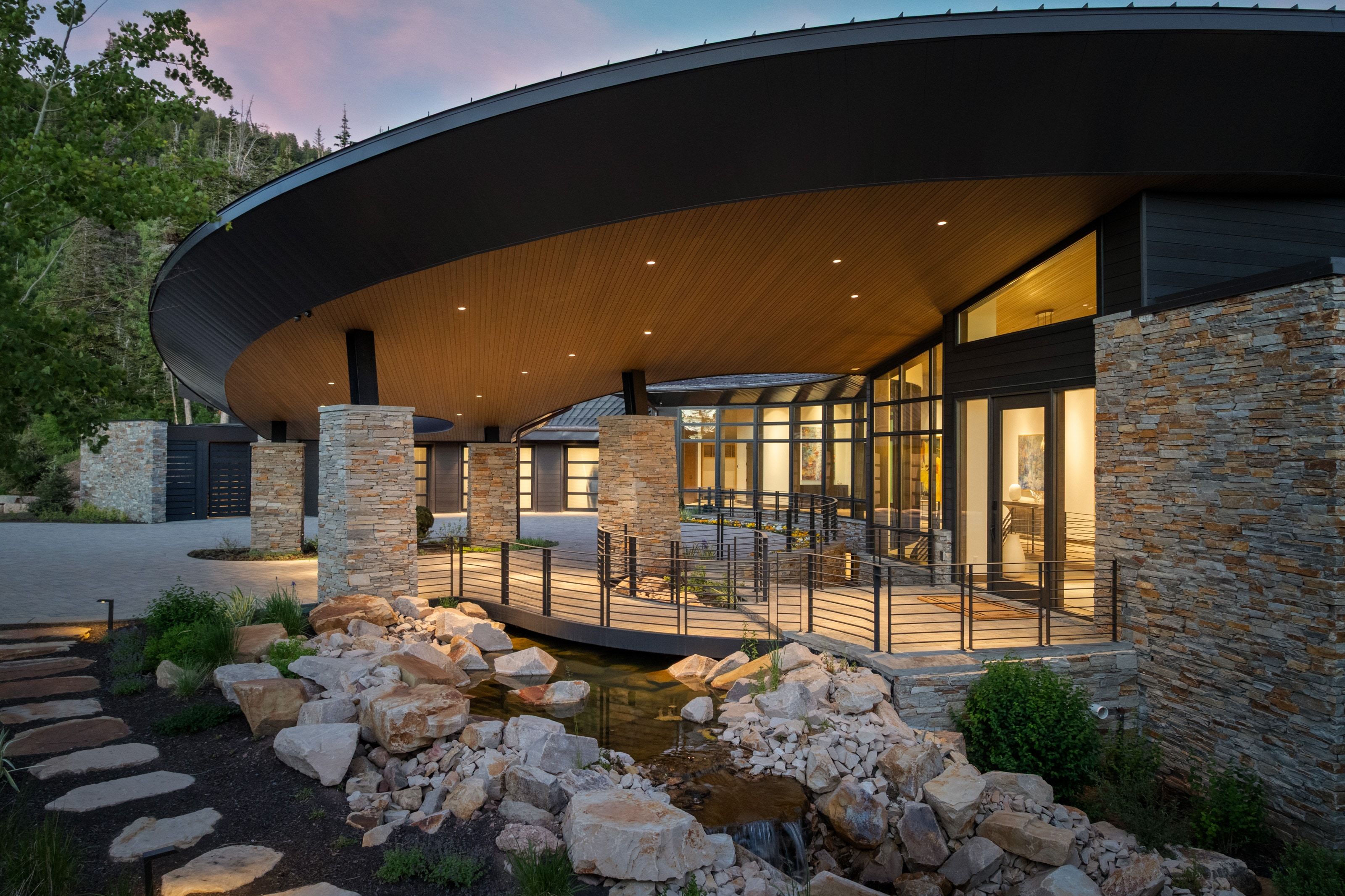 Contemporary exterior featuring a curved roofline with wood-finish soffit, stone veneer columns, and extensive glass walls