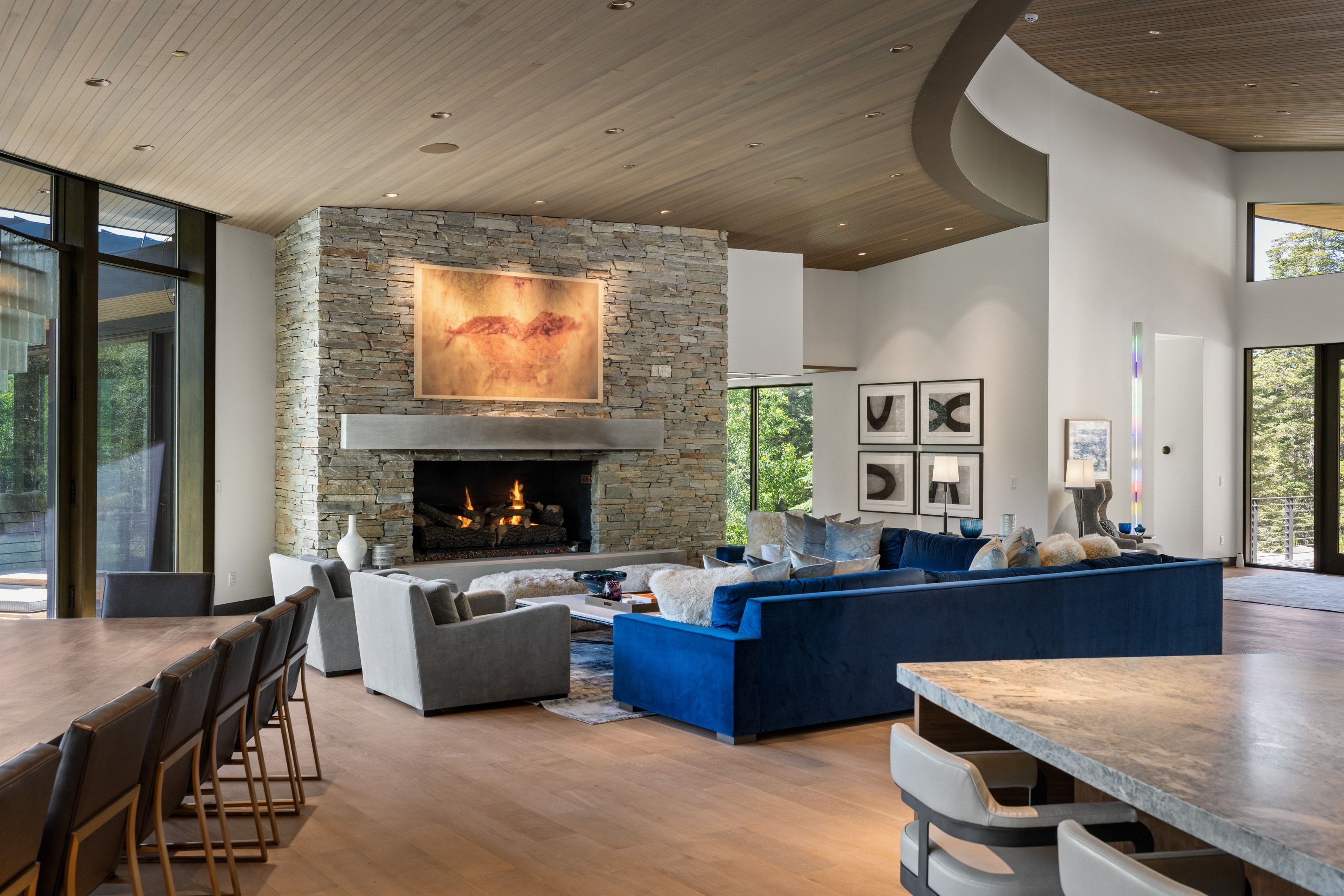 Spacious living area featuring a stone-clad fireplace with a concrete mantel, wood-finish flooring, and a light wood-paneled ceiling with recessed lighting