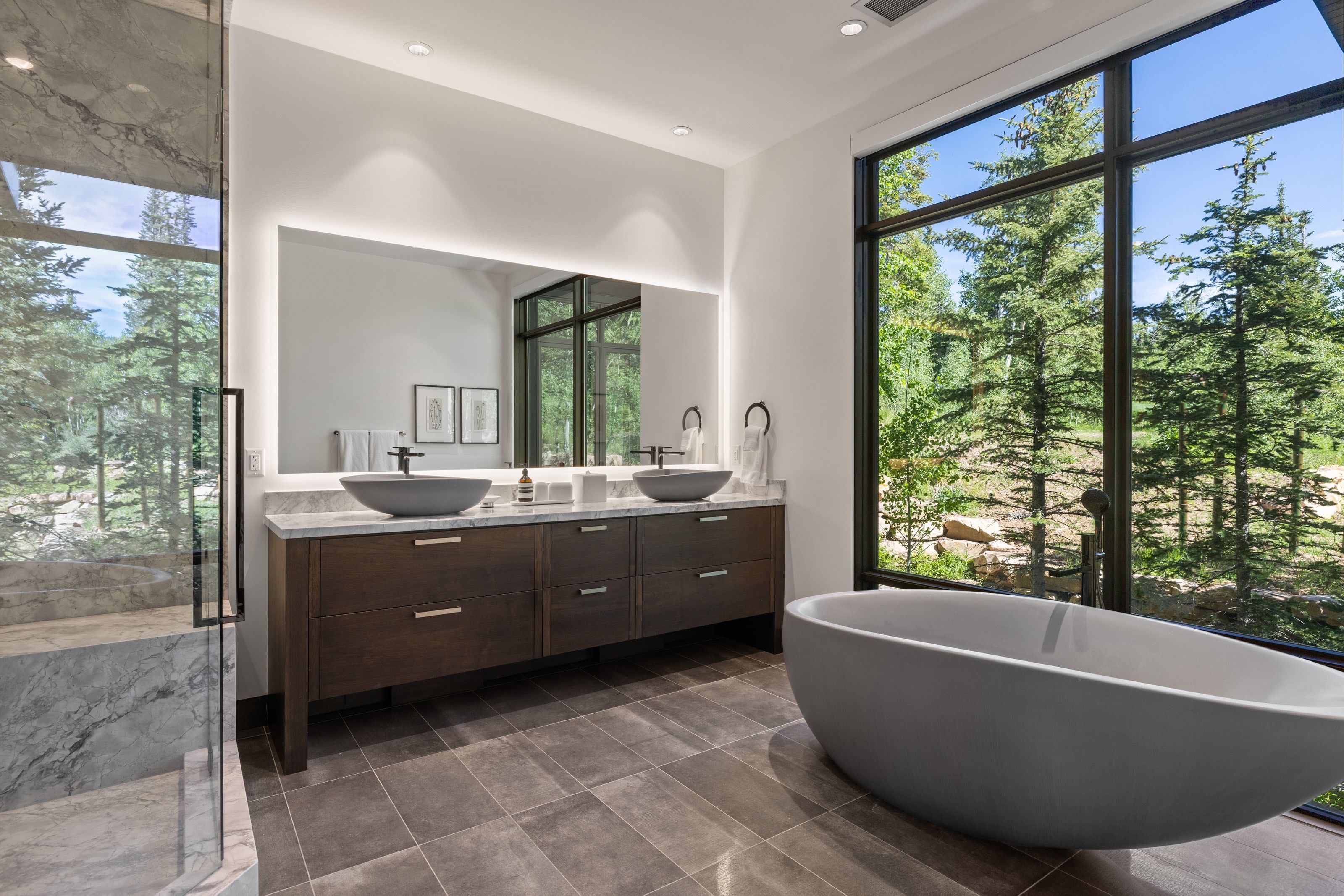 Spacious bathroom featuring a freestanding soaking tub, a large picture window, a dual vanity with vessel sinks, a backlit mirror, and a glass-enclosed shower with stone-finish walls
