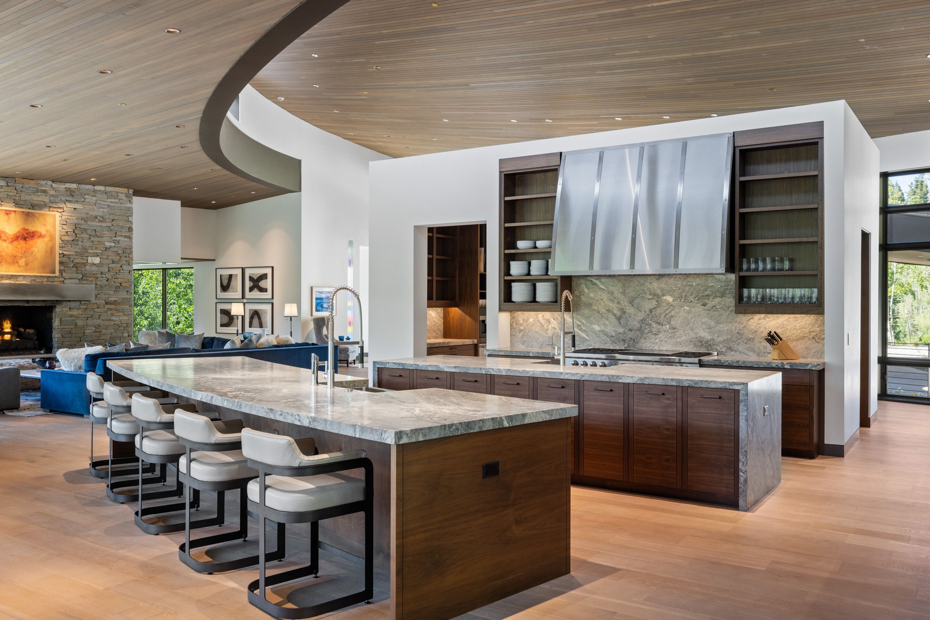 Open-concept kitchen featuring two large islands with stone countertops, integrated cooktop, a prominent stainless steel range hood, and rich wood-finish cabinetry