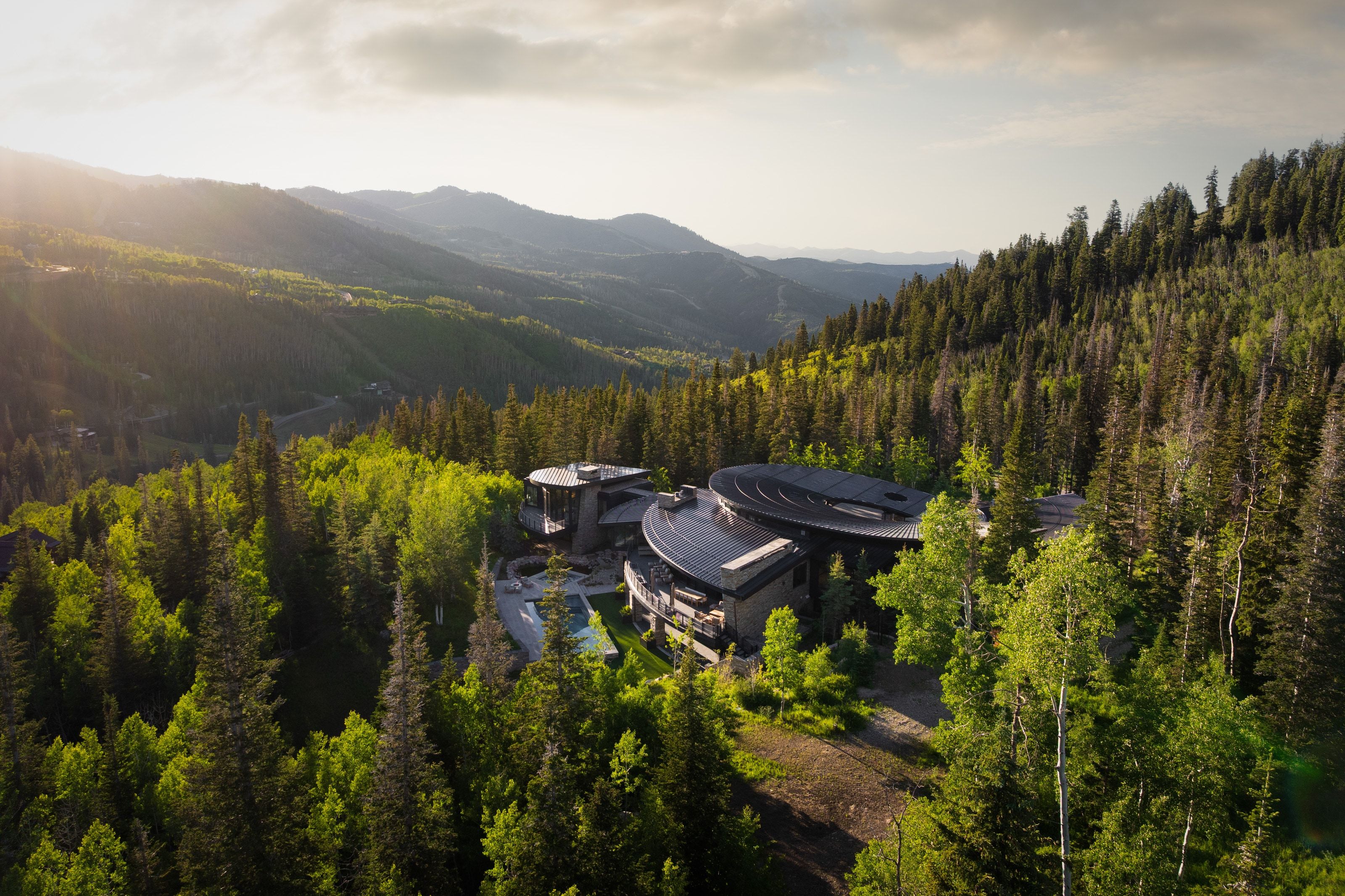 Architectural mountain residence nestled among dense evergreen forests, featuring a curvilinear roofline, expansive exterior decks, and an outdoor pool area