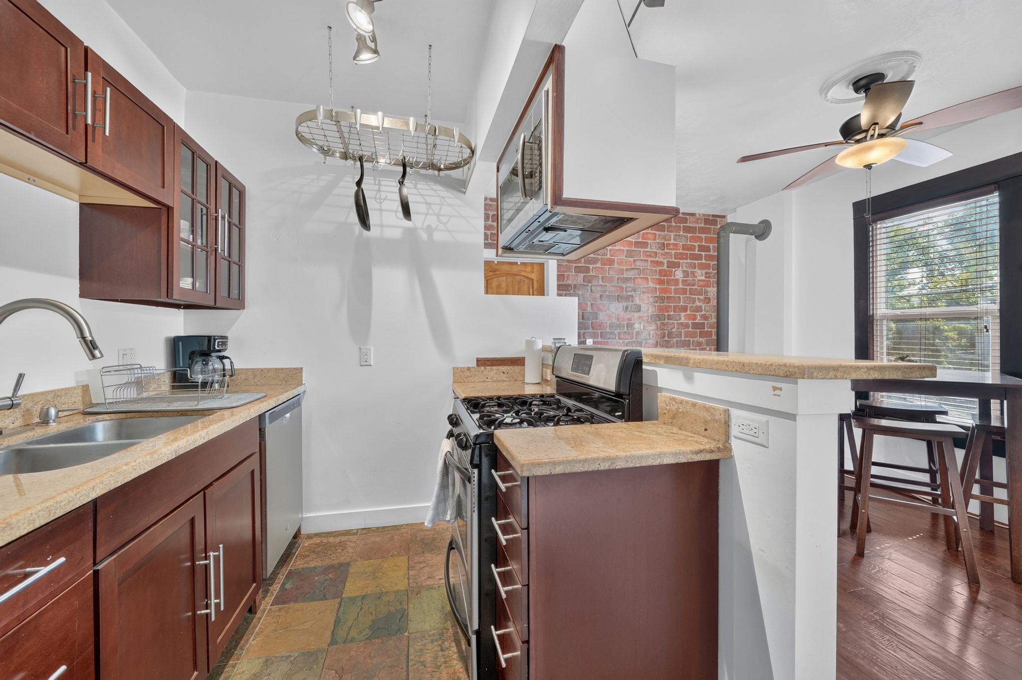Kitchen featuring rich wood cabinetry with glass-front accents, light-toned countertops, a stainless steel double basin sink with a gooseneck faucet, a pot rack, and multi-toned slate-look flooring