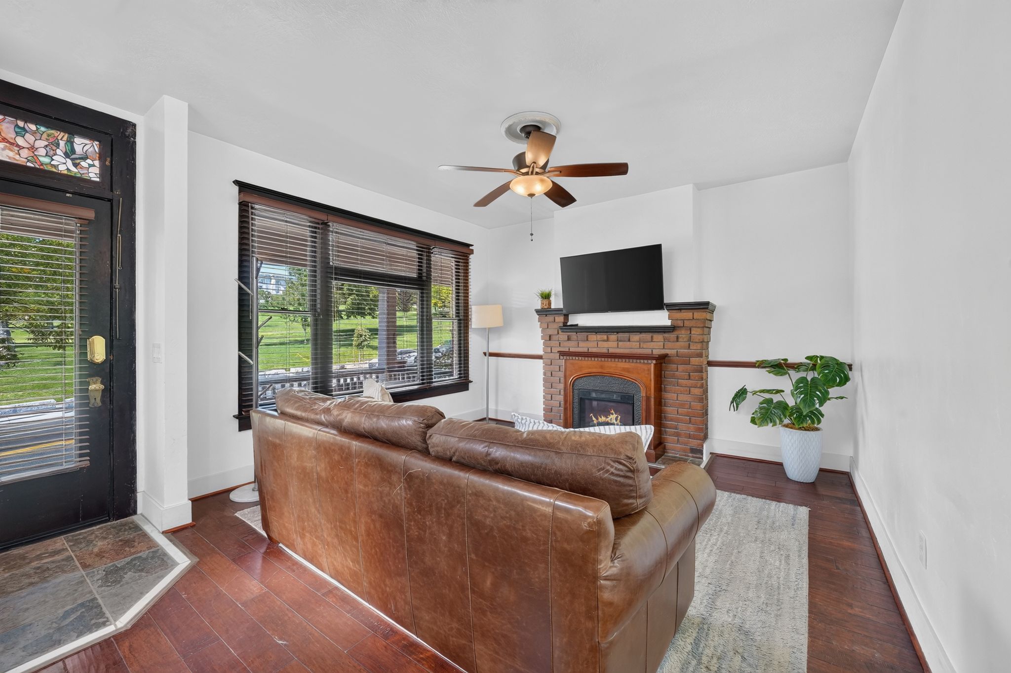 Living area featuring laminate flooring, a brick fireplace with a wood mantel, a ceiling fan with light fixture, and multiple windows