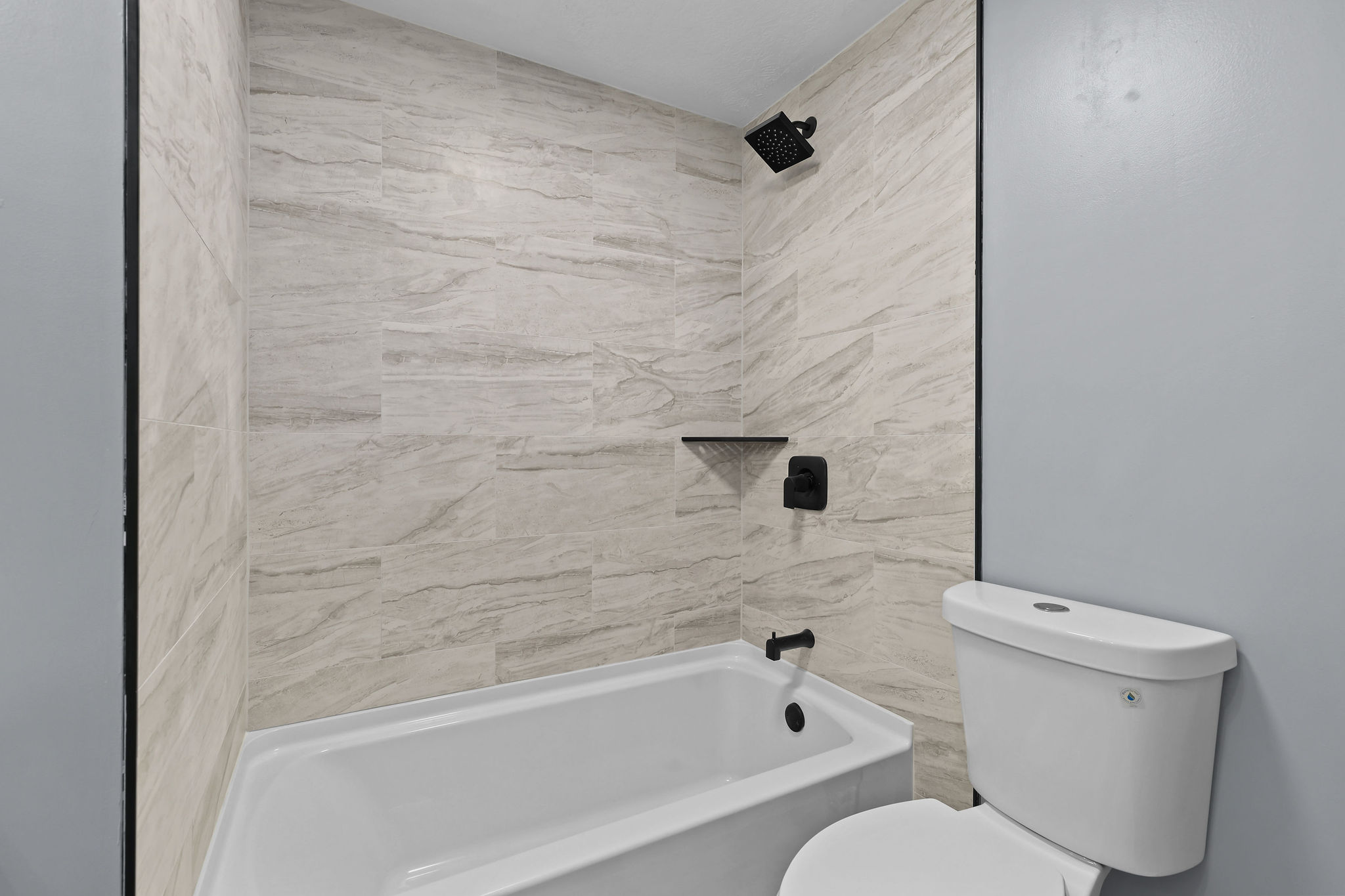 Bathtub and shower combination featuring large format stone-look tile surround, matte black shower fixtures, a built-in shower shelf, and a white porcelain tub