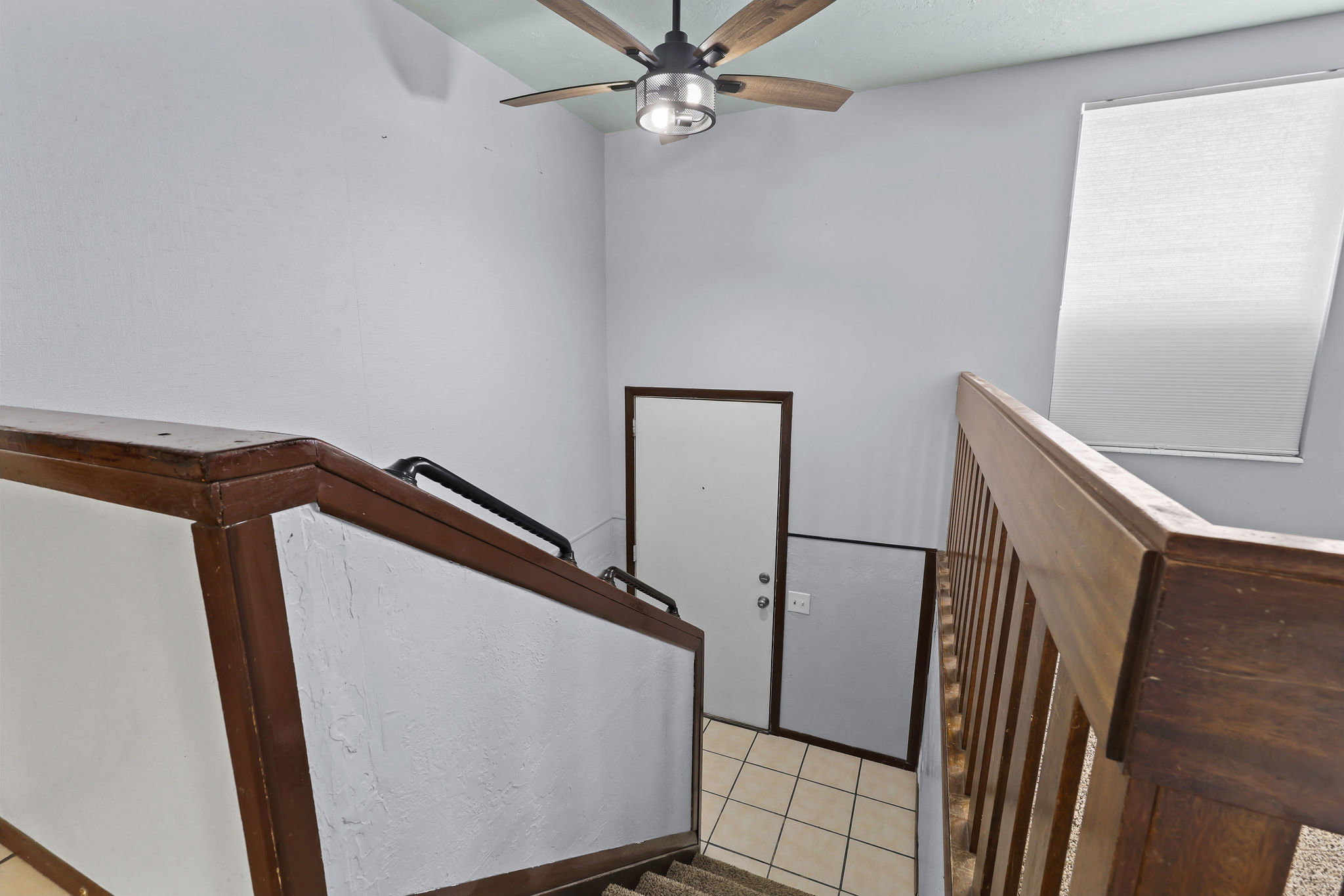 Stairwell with wood-finish railings, a ceiling fan with light fixture, and a window with a roller shade
