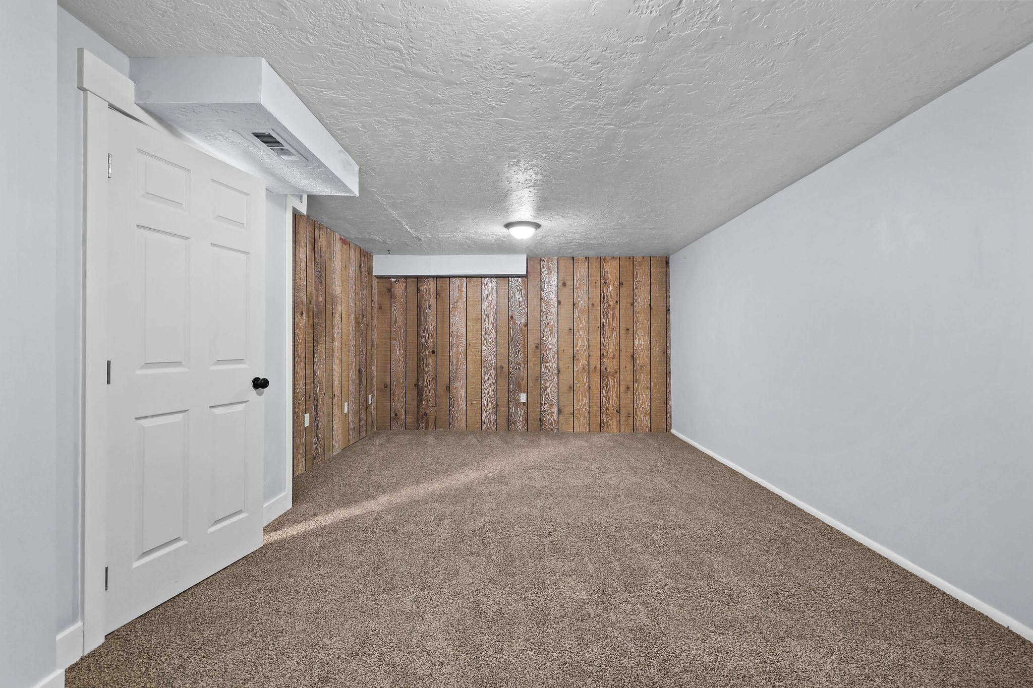 Carpeted room featuring a white paneled door with black hardware, a wood-paneled accent wall, light grey painted walls, and a recessed ceiling light fixture