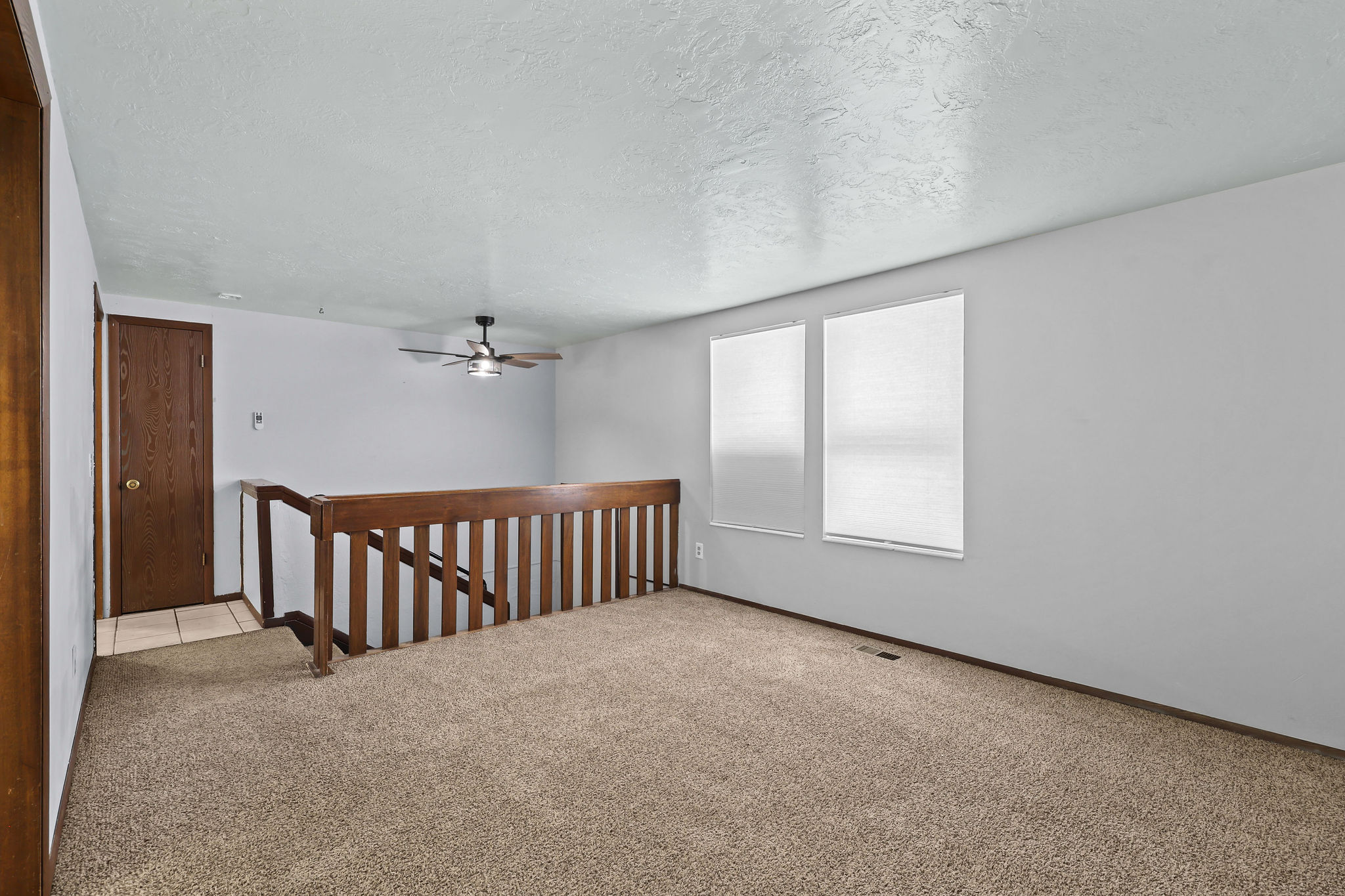 Carpeted living space with a ceiling fan, two windows, and a wood railing overlooking a staircase