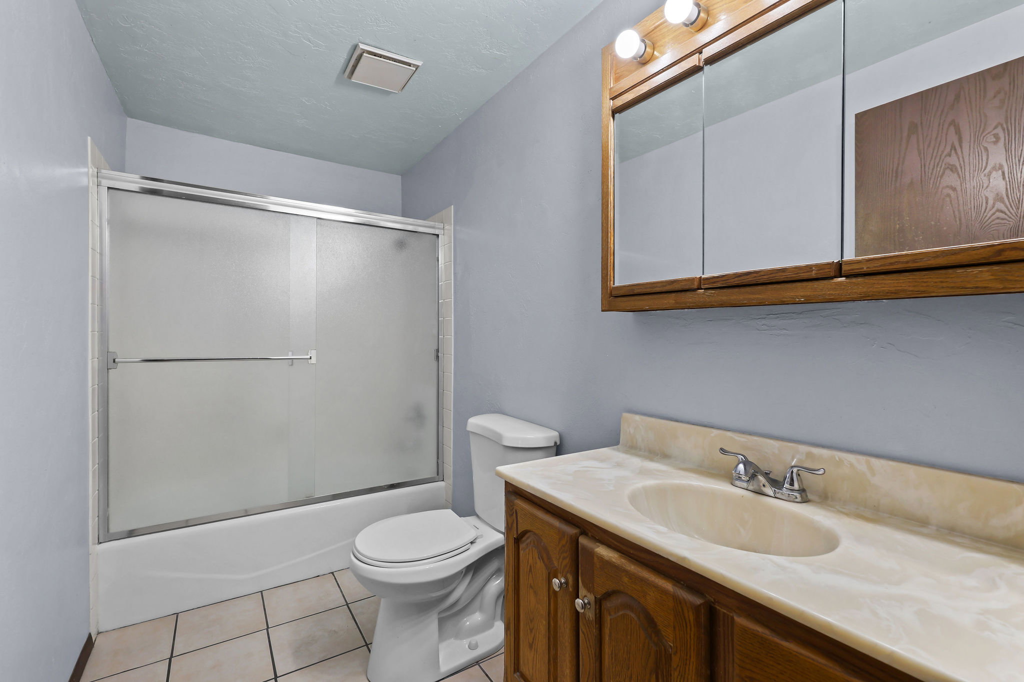 Bathroom featuring a built-in tub with sliding frosted glass doors, a vanity with wood-finish cabinetry, an integrated sink, and a mirrored medicine cabinet with overhead lighting