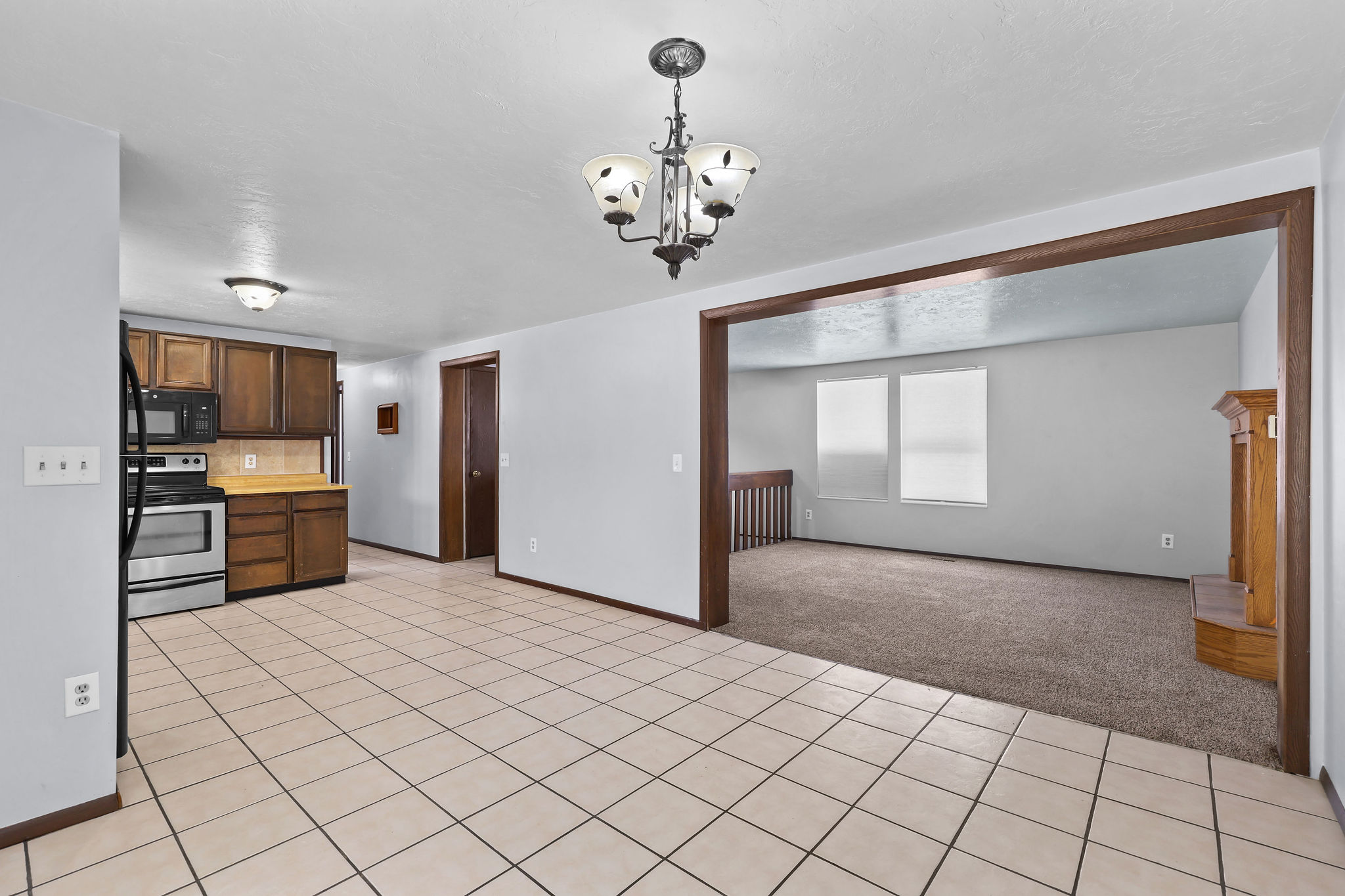 Spacious open-concept layout featuring a kitchen with wood cabinetry and stainless steel appliances, a dining area with tile flooring and an overhead chandelier, and a carpeted living space with a decorative wood mantel