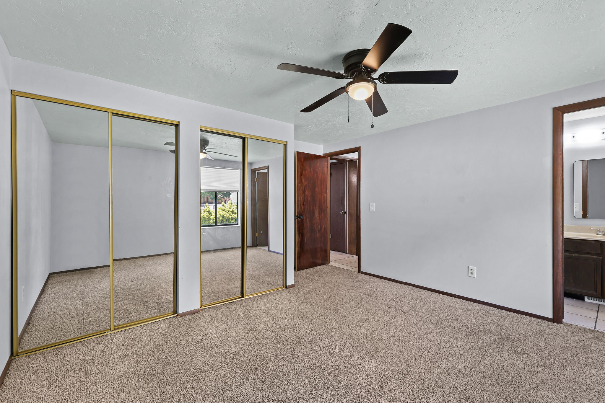 Carpeted interior space featuring a ceiling fan with light fixture, mirrored sliding closet doors with gold-tone framing, and wood-trim doorways