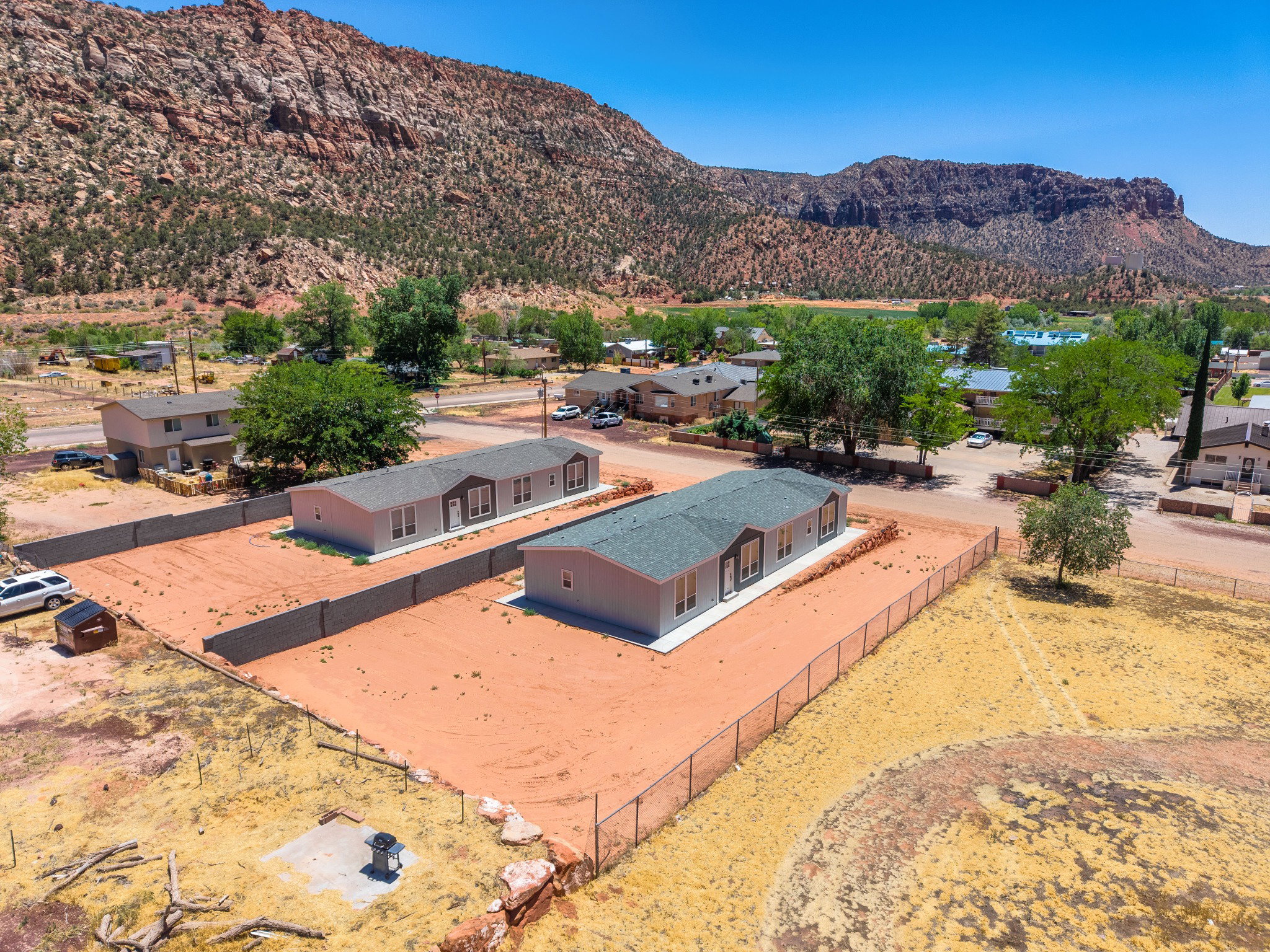 Two light gray modular homes with dark gray roofing, surrounded by red earth, feature multiple windows and front entryways