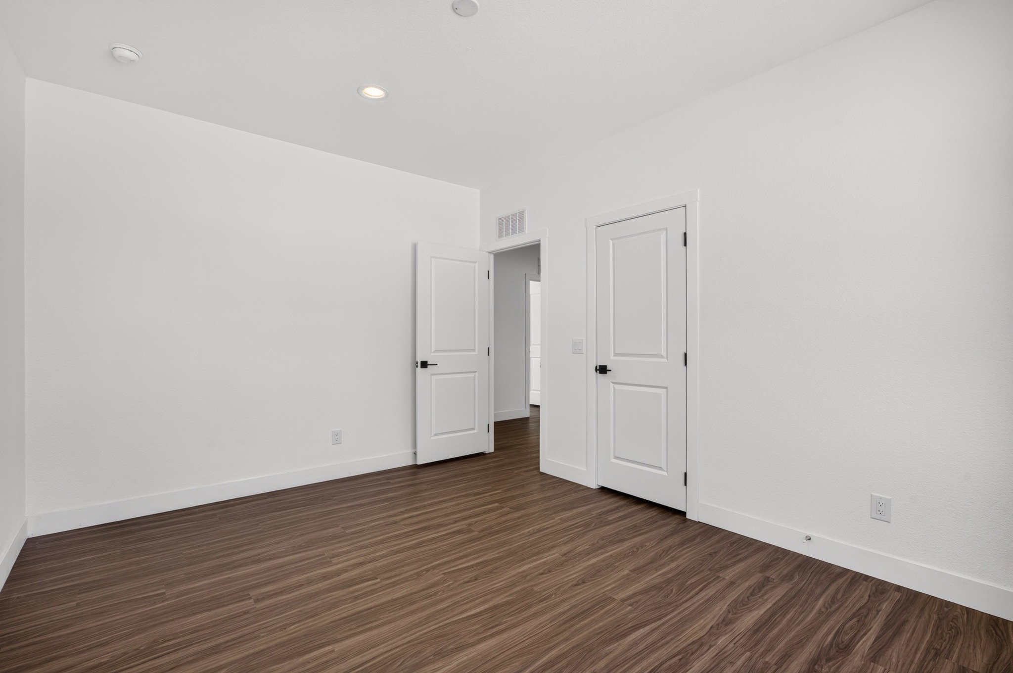 Spacious room featuring wood-finish flooring, white walls, recessed lighting, and two panel doors with matte black hardware