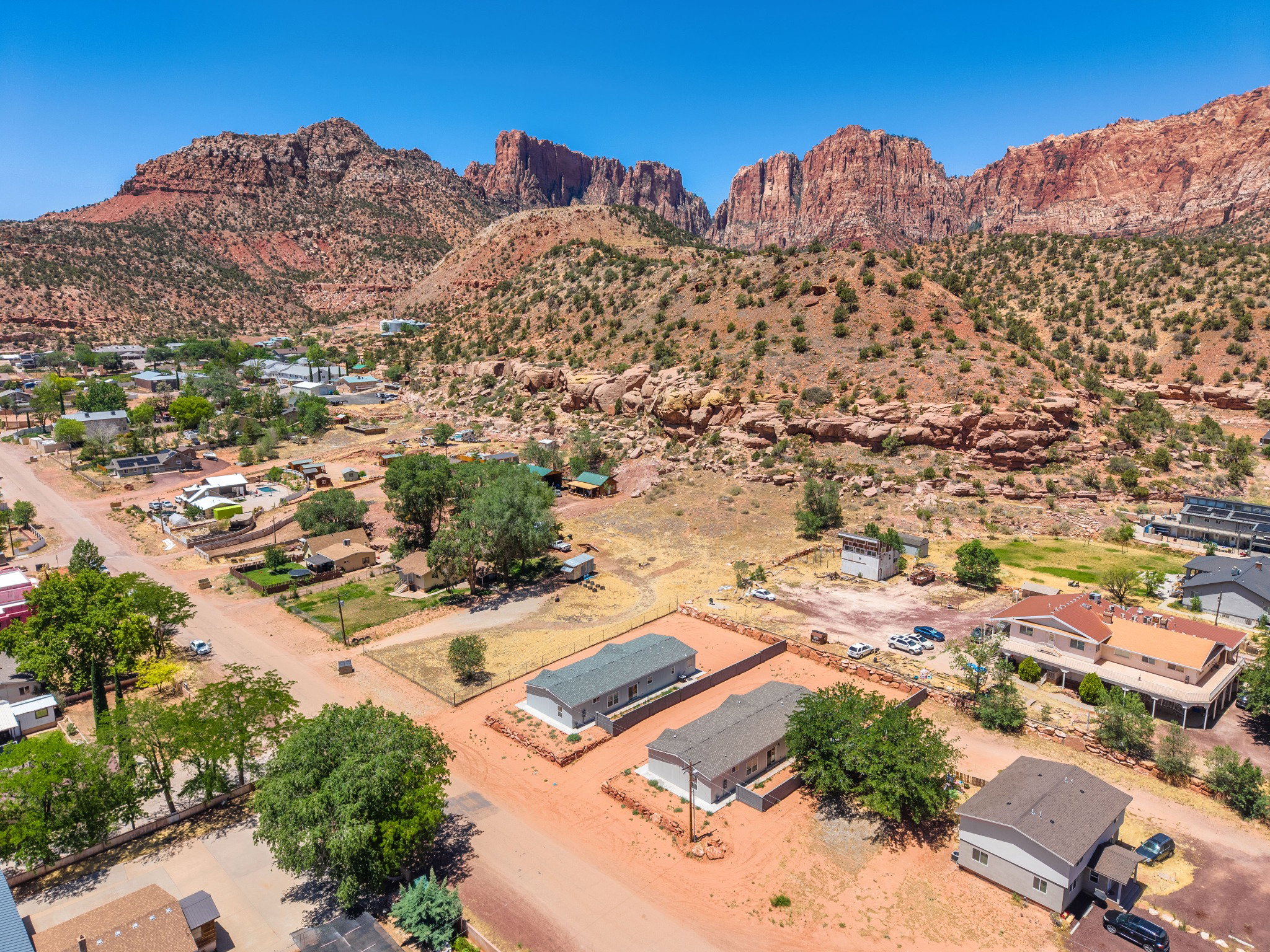 Expansive arid landscape featuring scattered residential properties, mature trees, unpaved roads, and prominent red rock formations in the background
