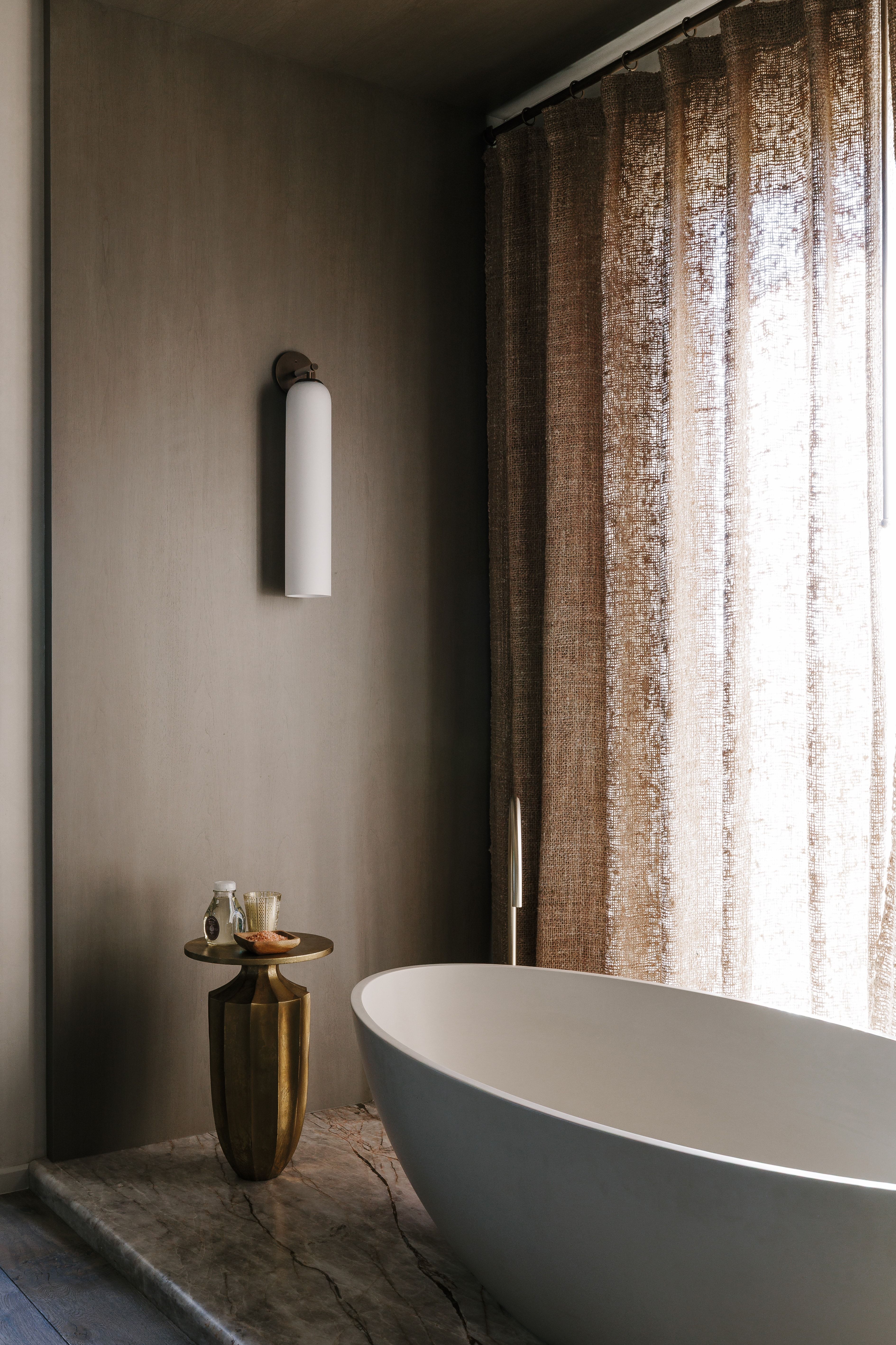 Freestanding soaking tub set on a stone slab, featuring textured wall finishes, a modern wall sconce, and natural light with full-length drapery