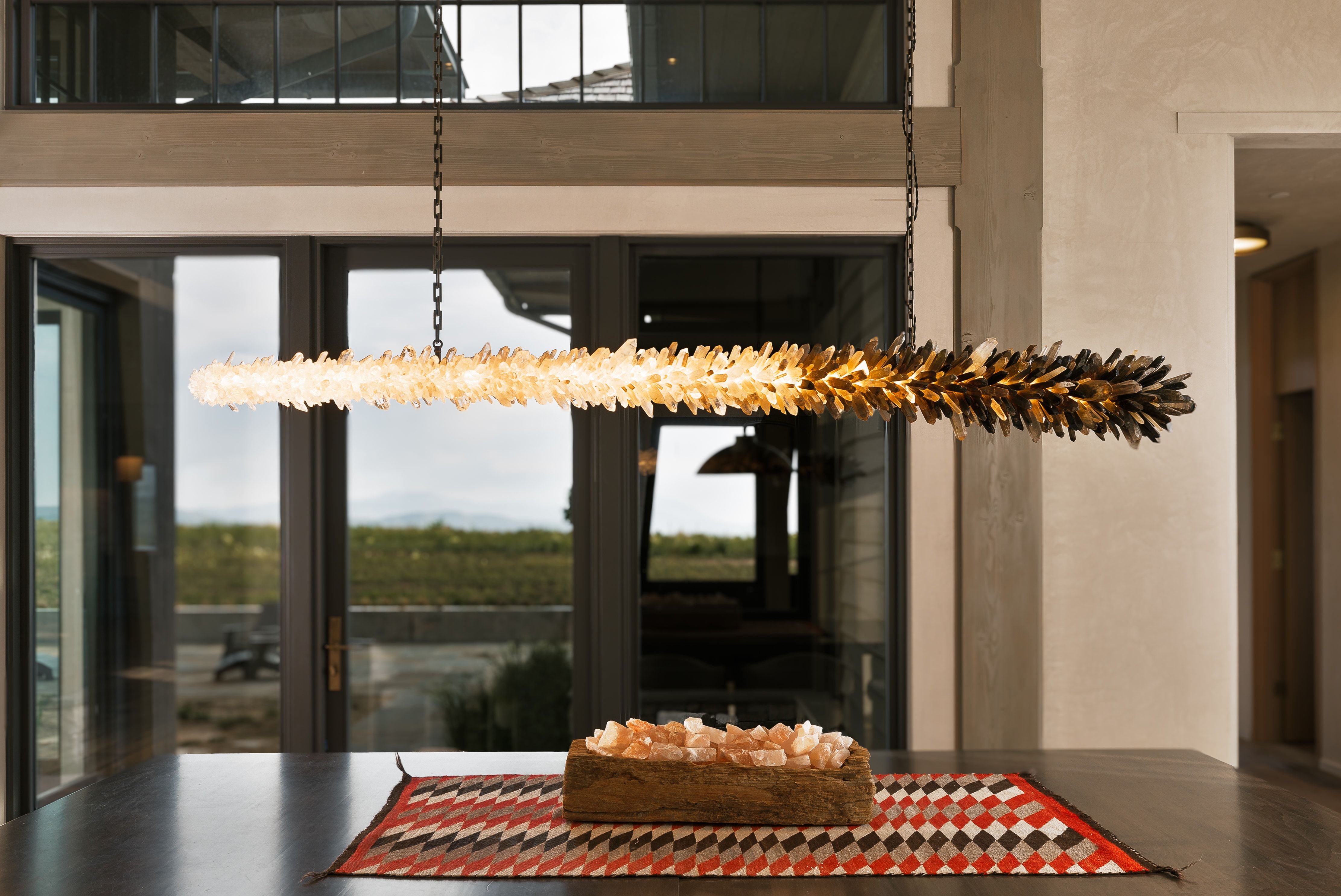 Dining area featuring a linear crystal chandelier, dark-toned dining table, and large glass doors opening to outdoor scenery