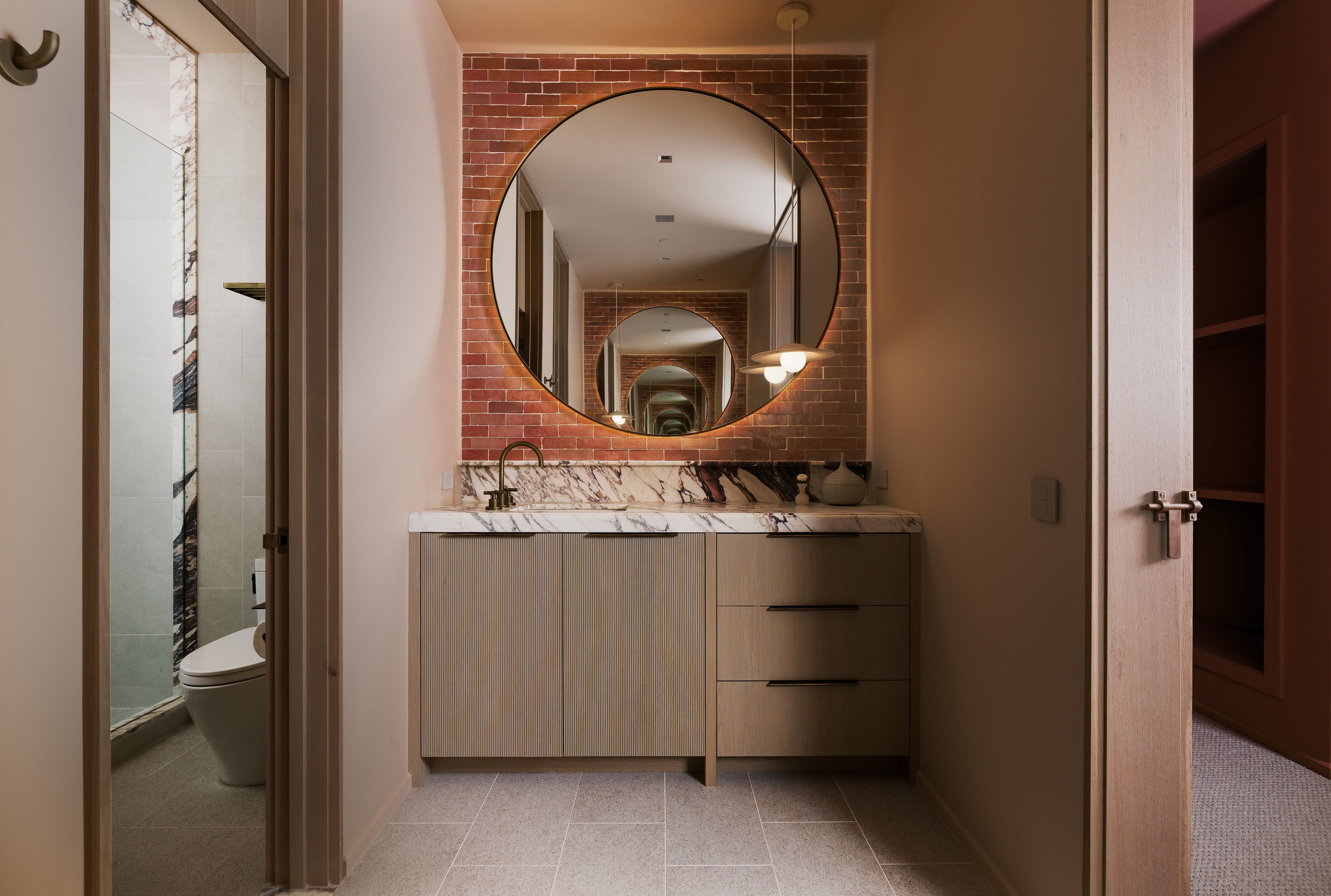 Contemporary vanity featuring a marble-patterned countertop, gold-finish faucet, and a large round mirror with integrated lighting