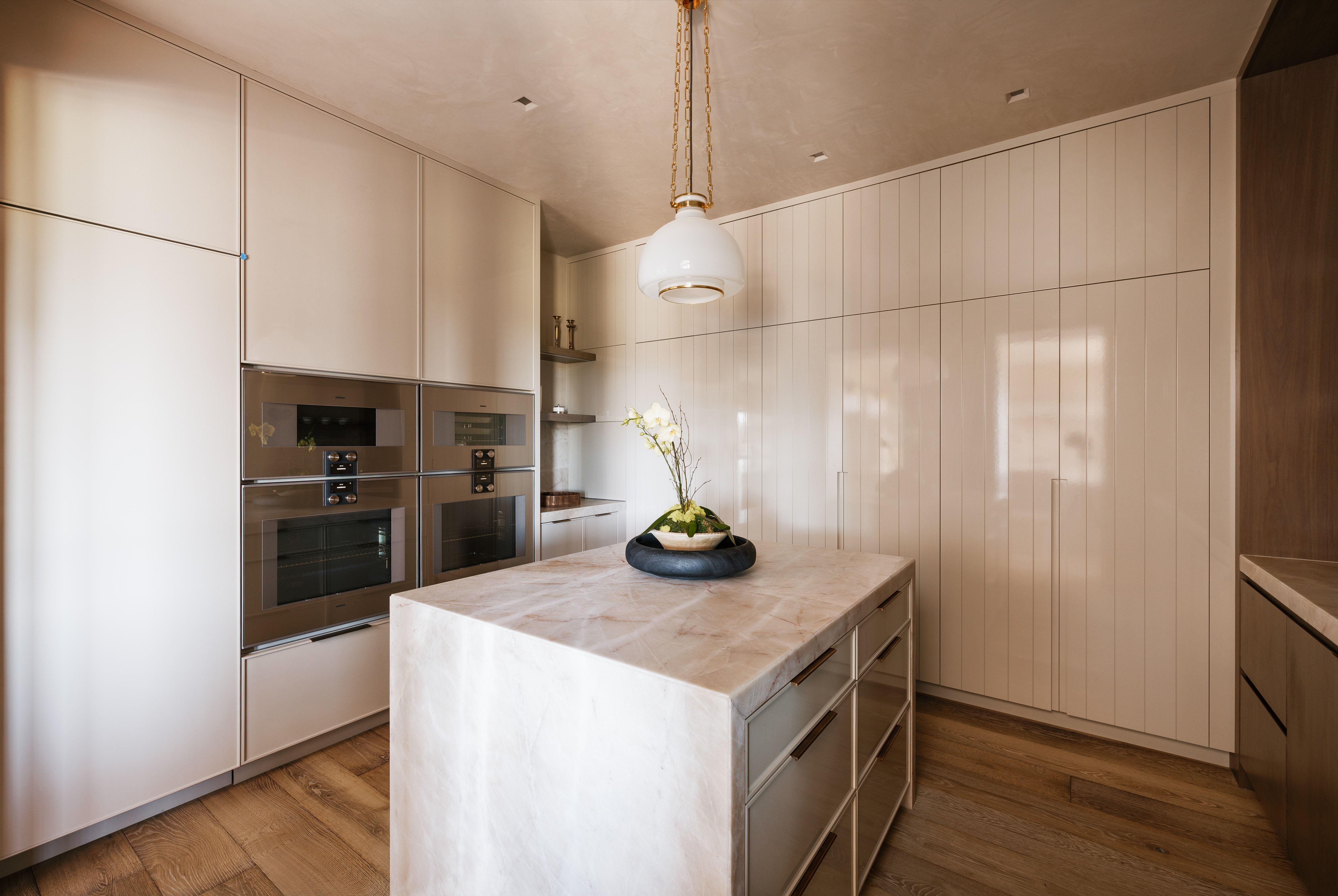 Gourmet kitchen featuring a waterfall island, integrated stainless-steel double ovens, custom cabinetry, wood-finish flooring, and a pendant light fixture