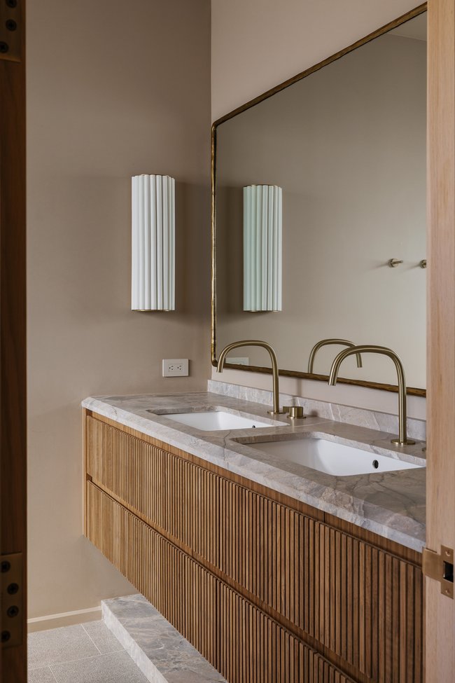 Floating wood-finish vanity featuring dual integrated sinks, stone countertop, and wall-mounted brass faucets