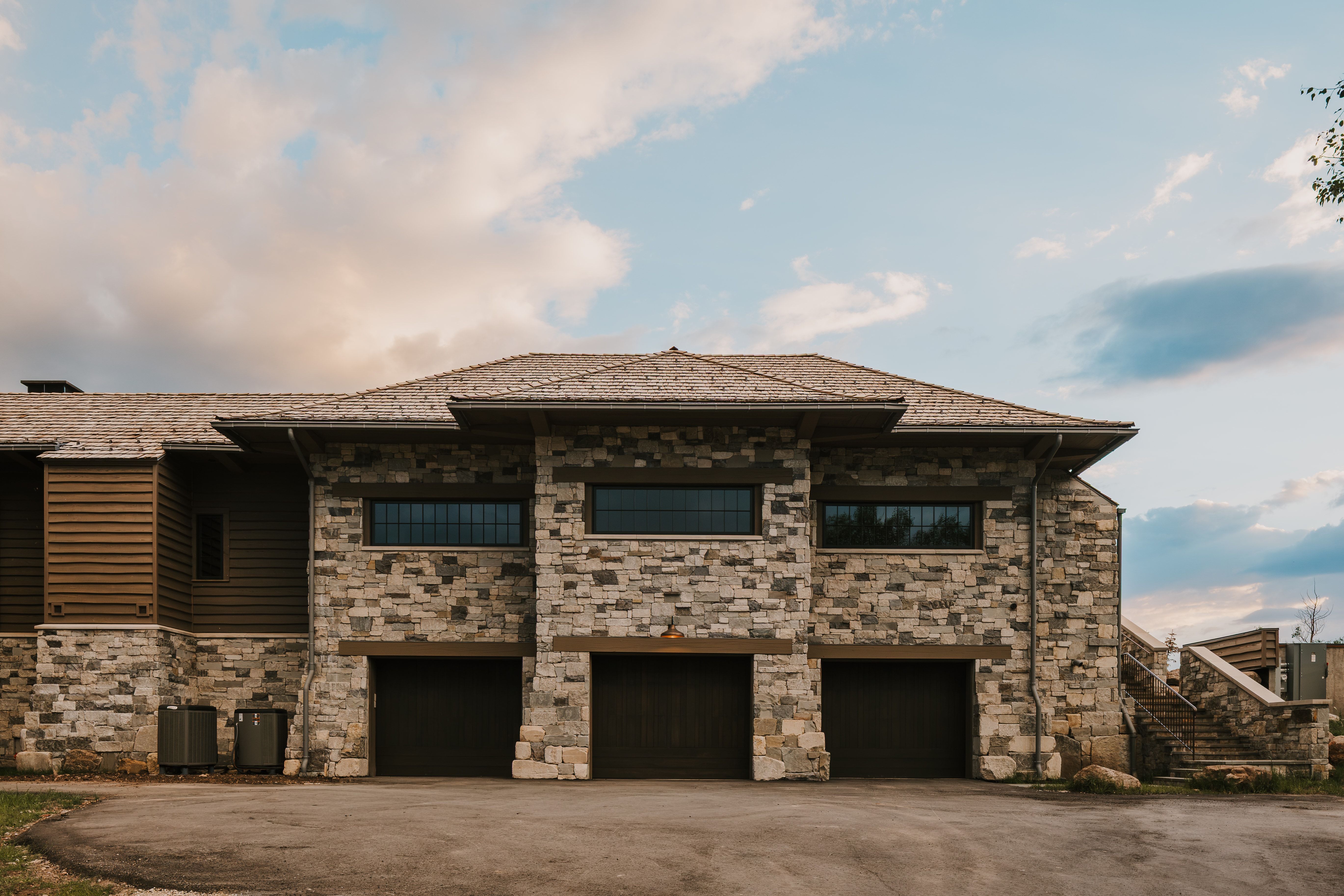Stone and wood-sided exterior featuring three garage bays, a shingle roof, and a stone staircase
