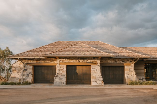 Three-car garage featuring natural stone facade, wood-finish garage doors, exposed wood beams, and a cedar shake roof