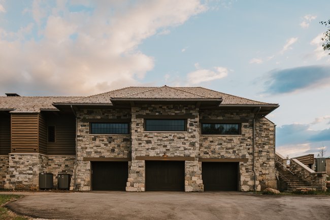 Stone and wood-sided exterior featuring three garage bays with dark doors, a shingle roof, and multiple upper-level windows
