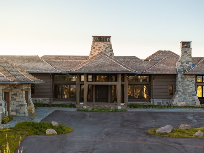 Exterior featuring natural stone accents, cedar shake roofing, multiple stone chimneys, expansive windows, and a paved driveway with stone detailing