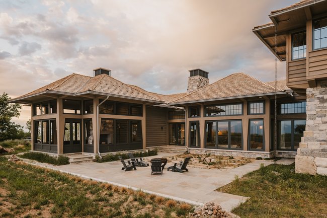 Expansive rear exterior featuring a stone patio, large picture windows, a stone-clad chimney, and a cedar shake roof