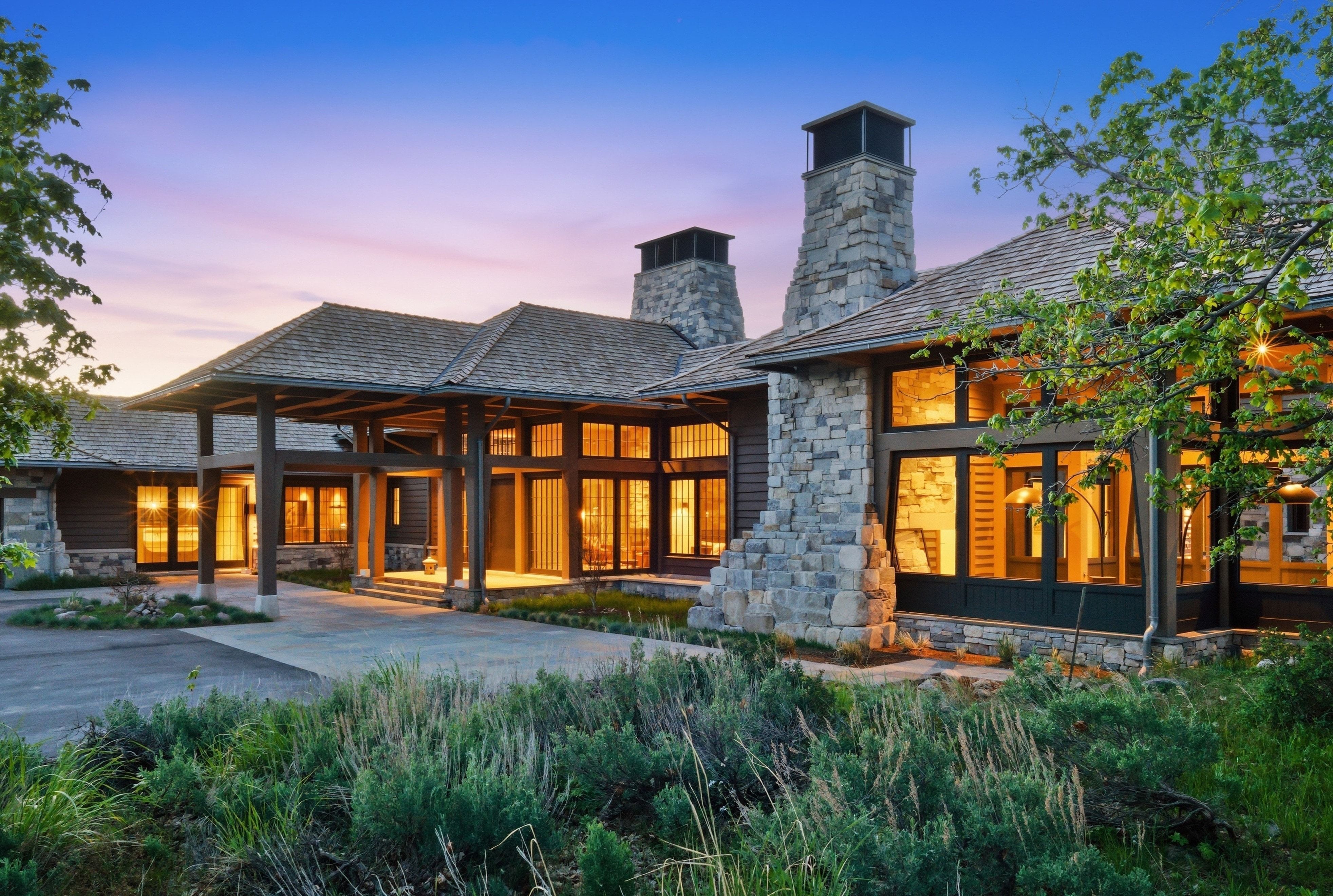 Stone and timber exterior featuring multiple large picture windows, a covered porte-cochère entrance, and two prominent stone chimneys