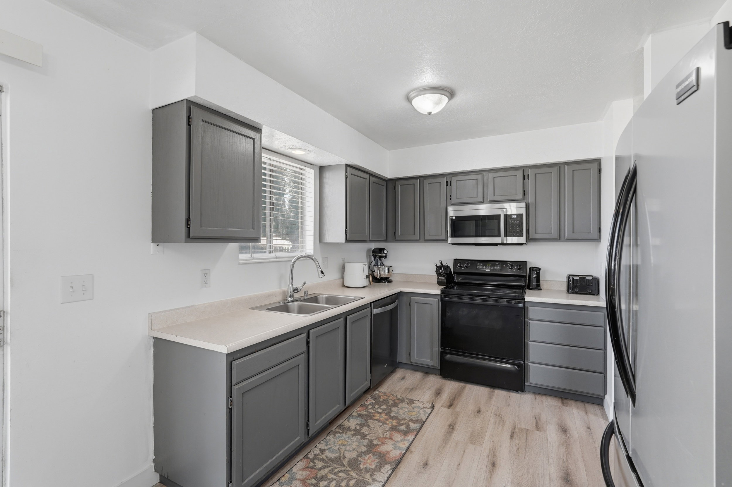 Kitchen featuring L-shaped gray cabinetry, a double basin stainless steel sink, light-toned countertops, and wood-finish flooring