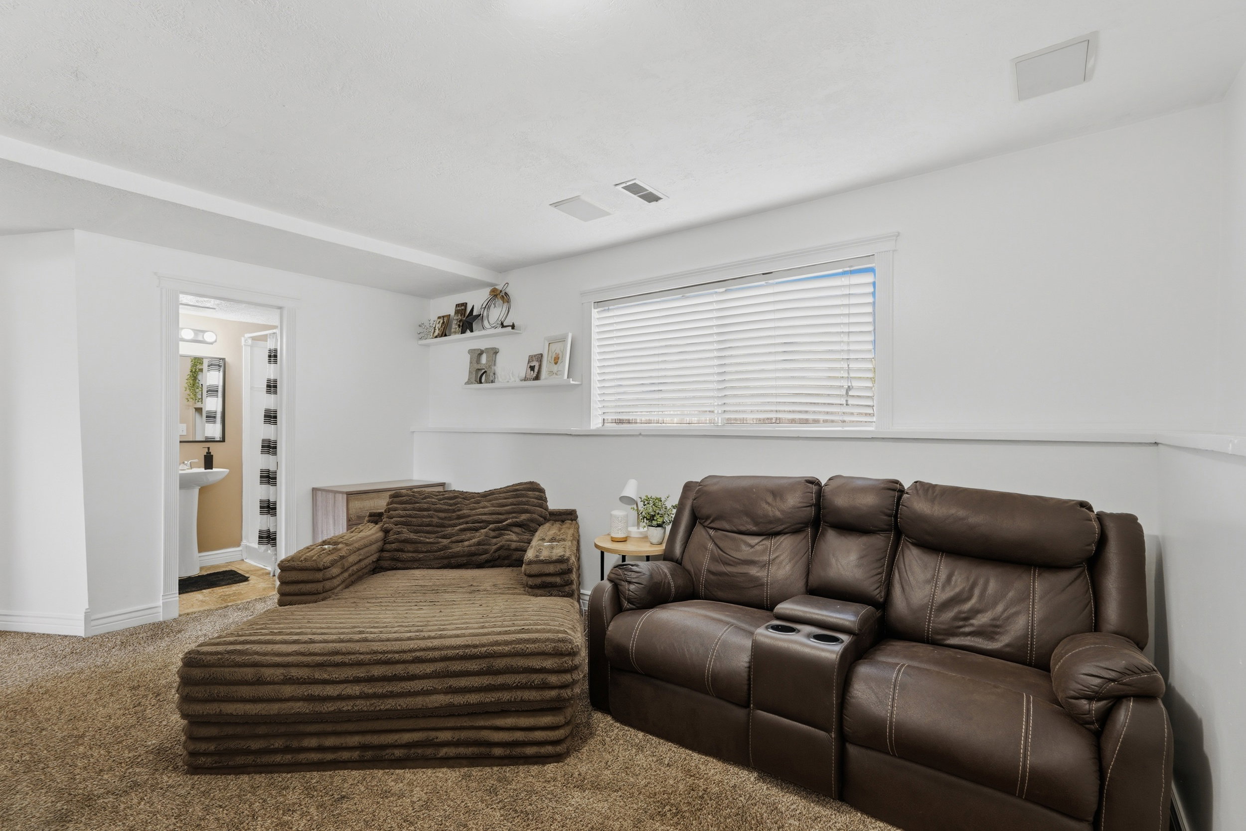 Spacious carpeted room featuring a window with blinds, white walls, and a visible doorway leading to a bathroom with a pedestal sink and vanity lighting