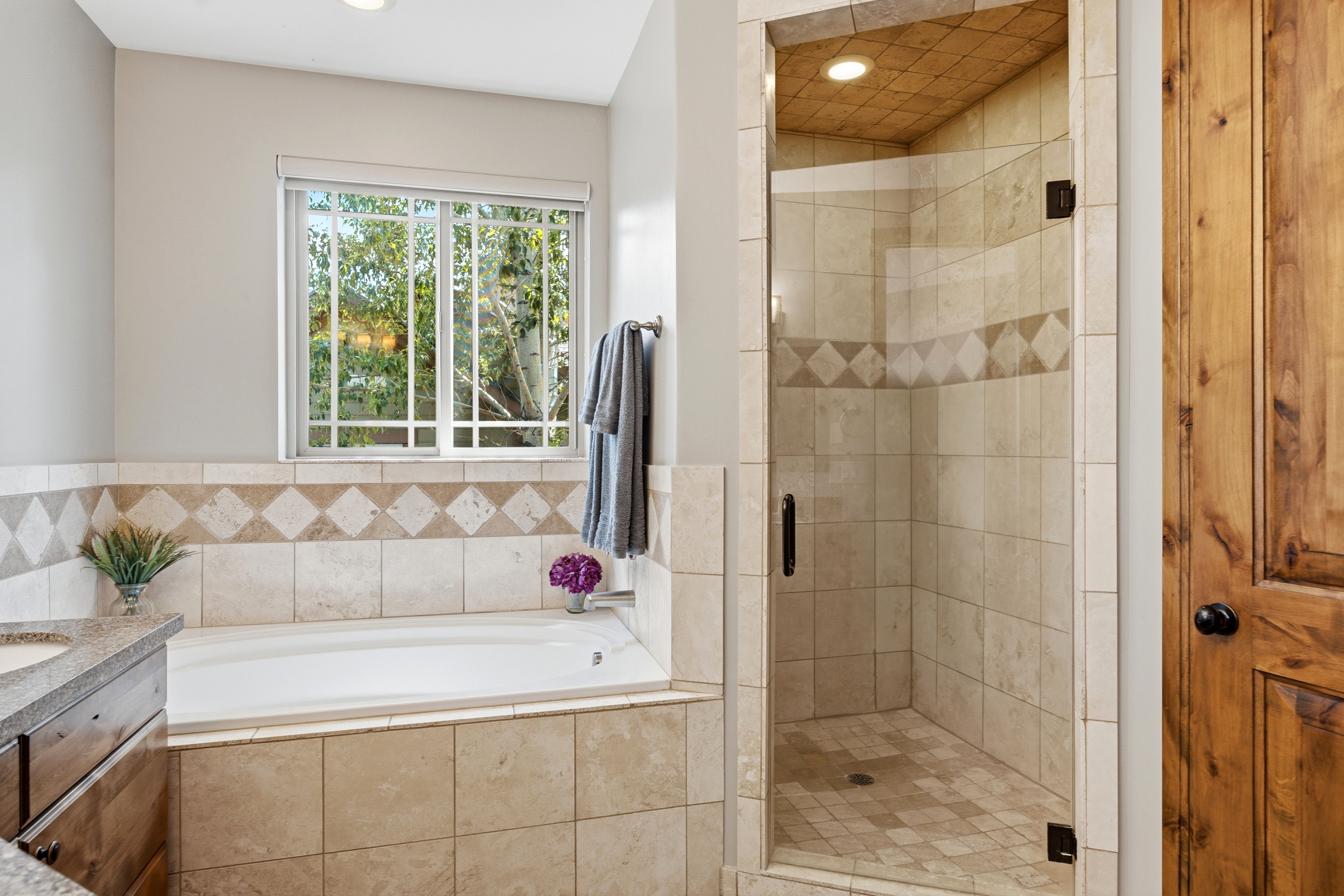 Bathroom featuring a built-in soaking tub with surrounding tile, a glass-enclosed shower with tile walls and a patterned accent band, a window with exterior tree scenery, wood-finish cabinetry, and a dark wood door