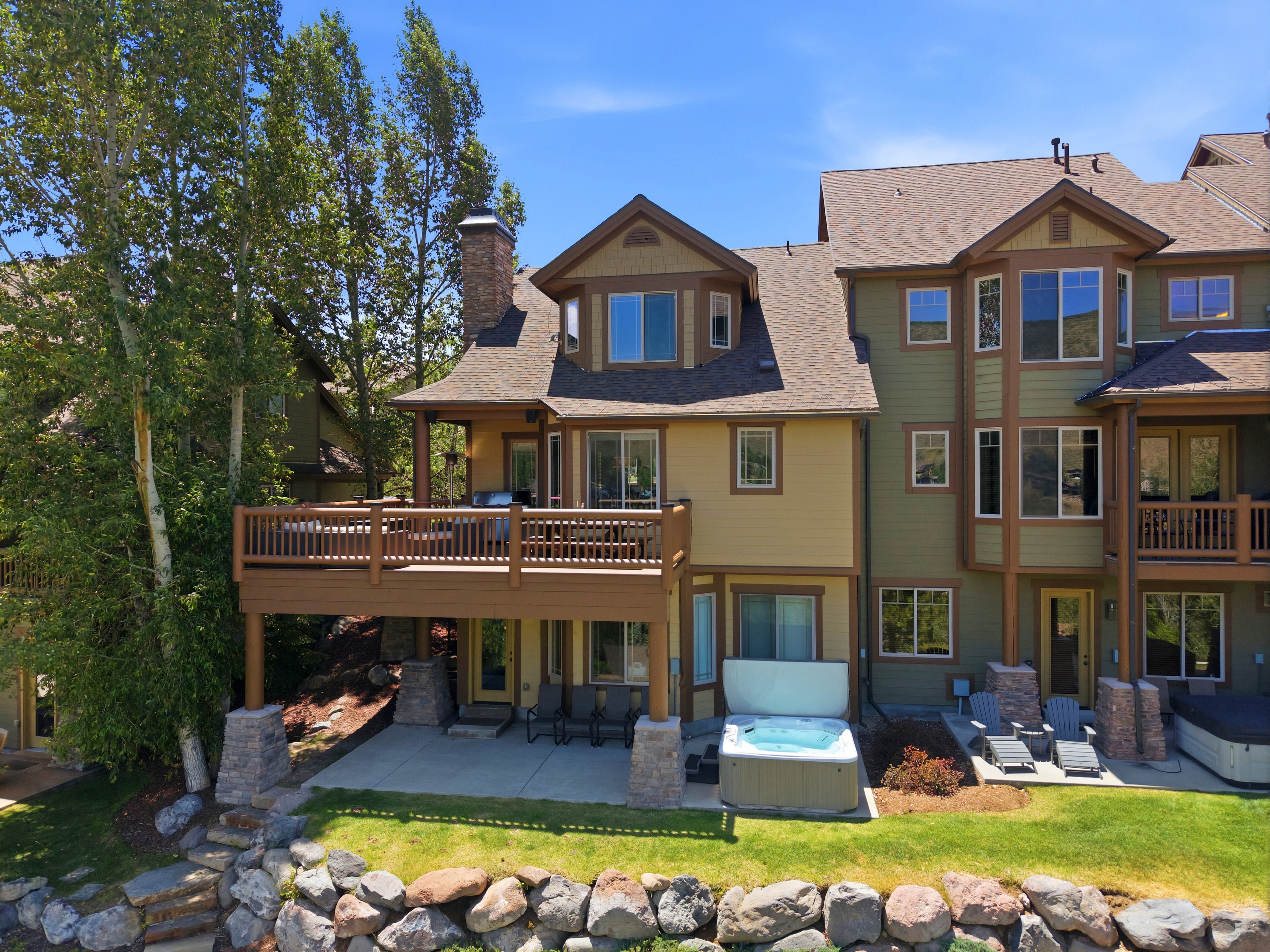 Multi-level townhouse exterior featuring a wood-finish deck, stone retaining wall, and a ground-level patio with a spa