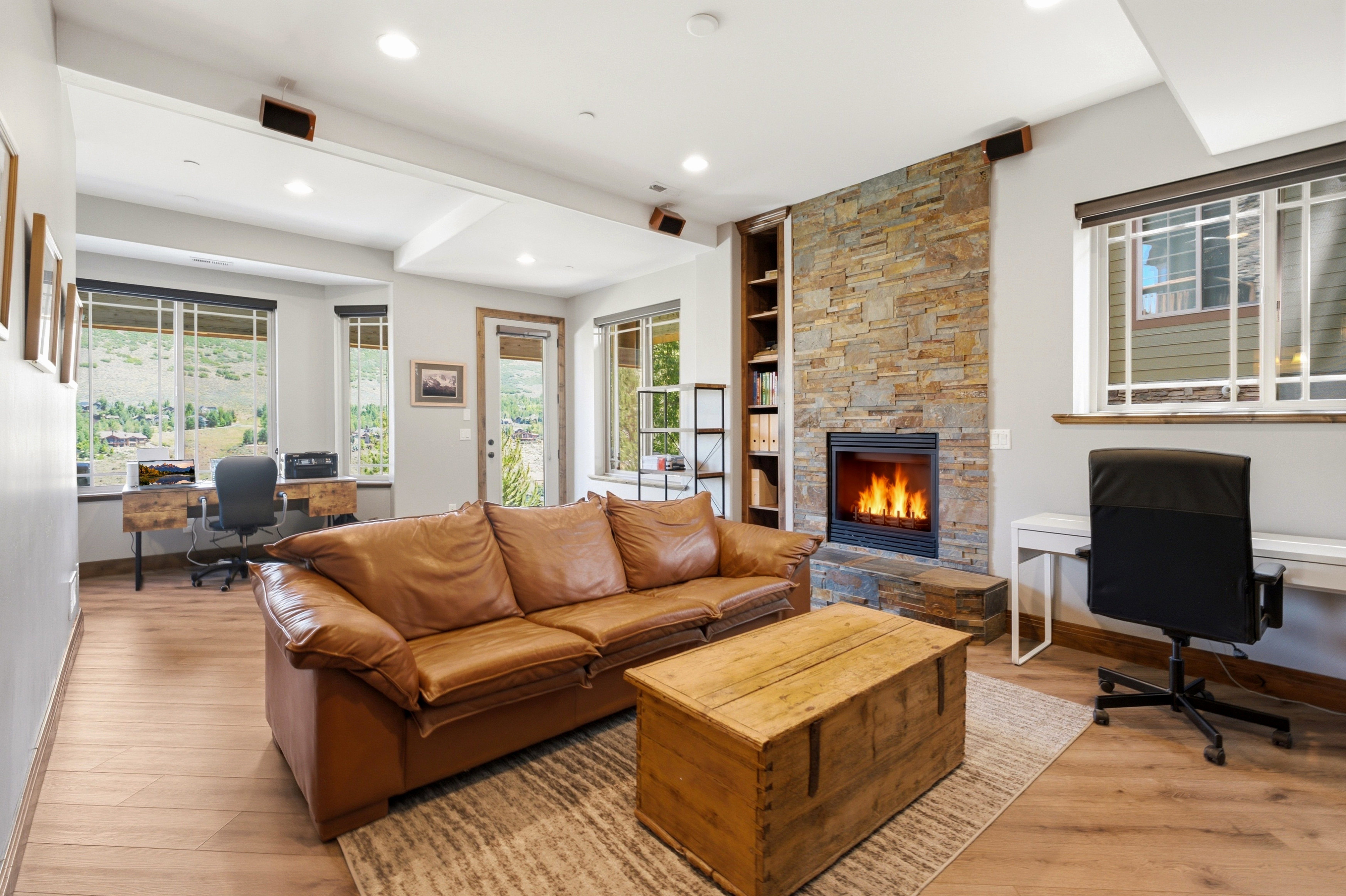 Open-concept living area featuring wood-finish flooring, a stone-clad fireplace with built-in shelving, recessed lighting, and multiple windows with mountain scenery
