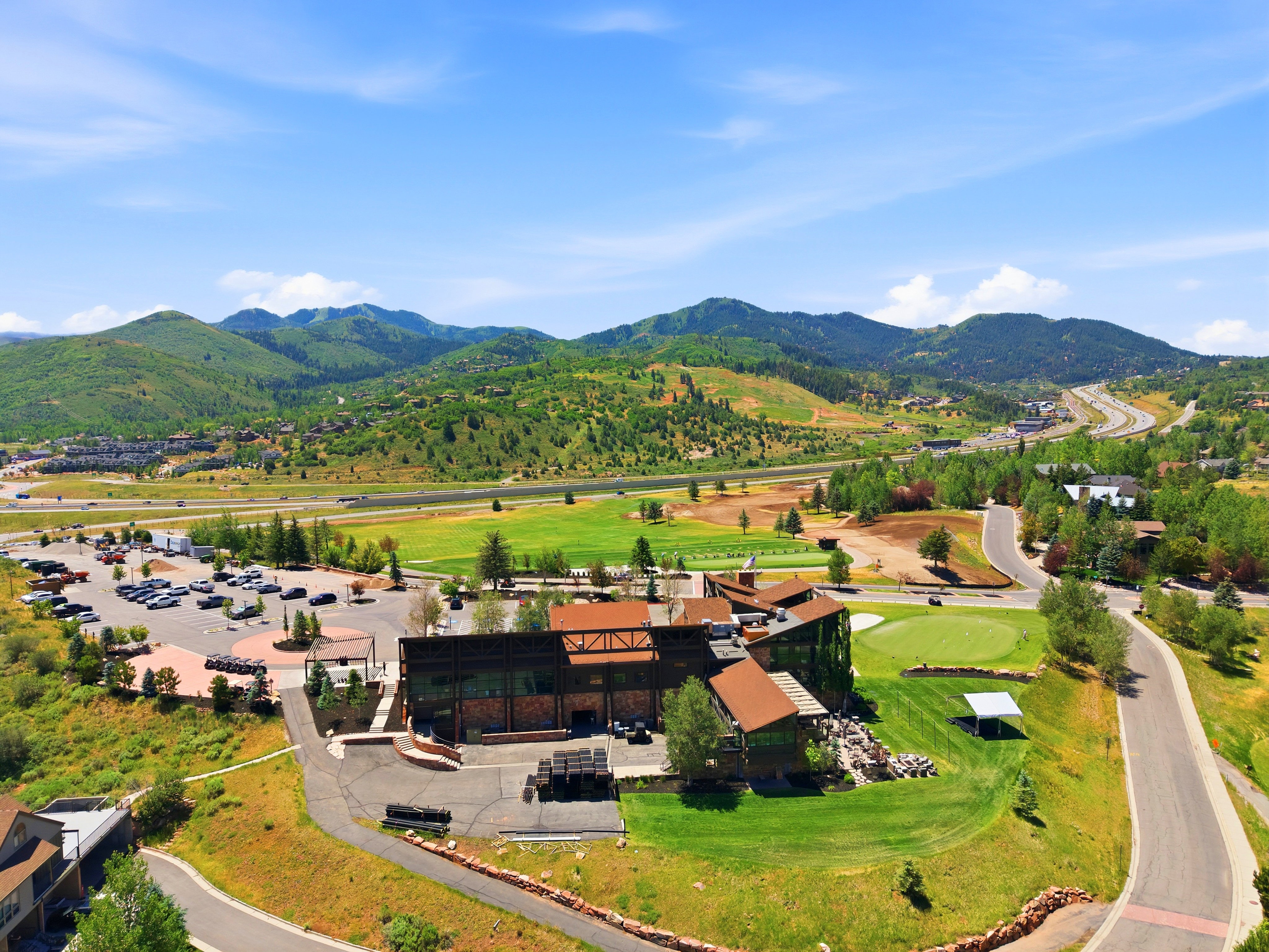 Mountainous landscape featuring a large commercial building with multiple rooflines, an expansive parking area, paved roads, and a manicured golf course green