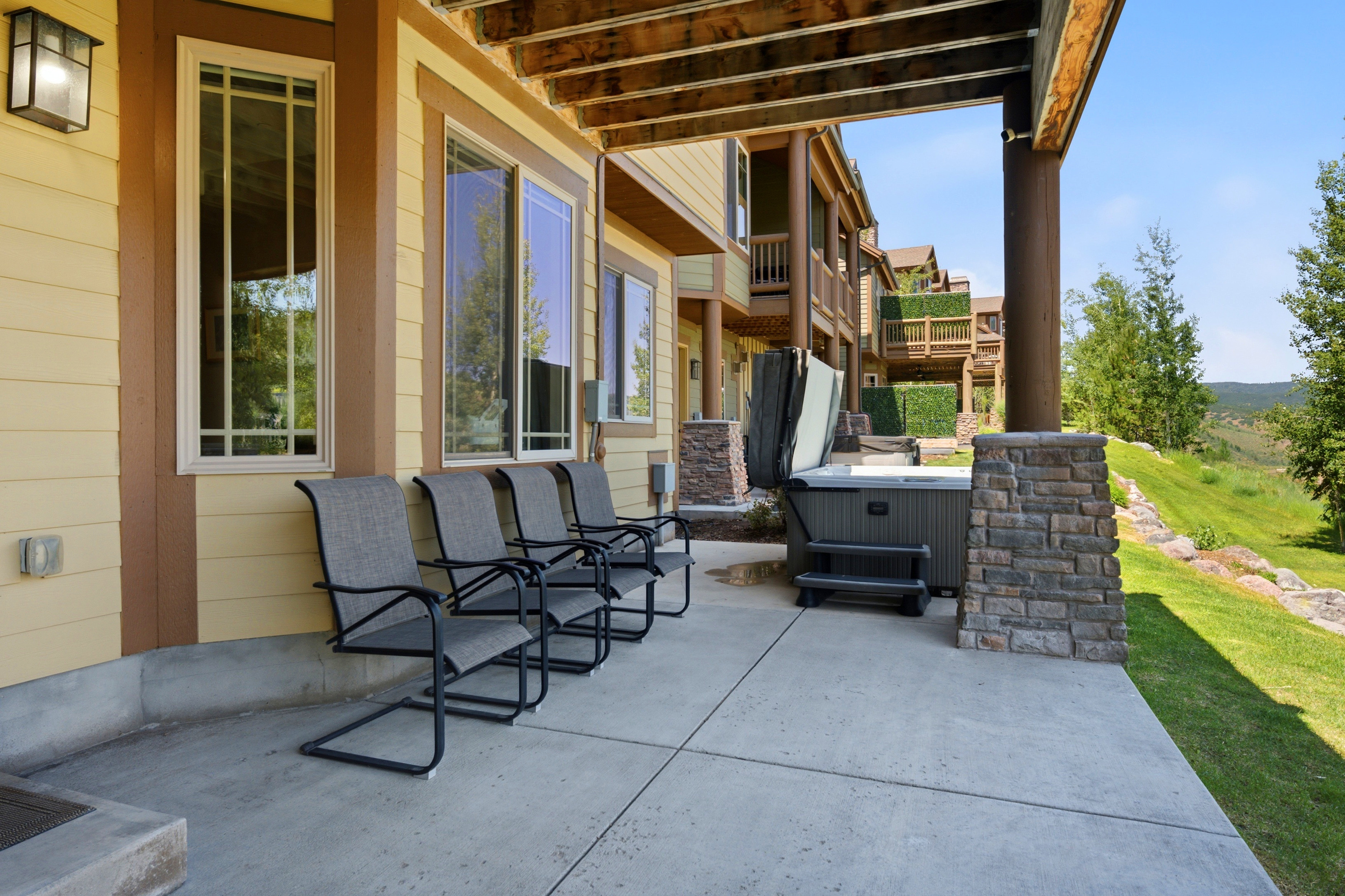 Covered concrete patio featuring a hot tub, stone-clad structural columns, and a landscaped grassy hillside