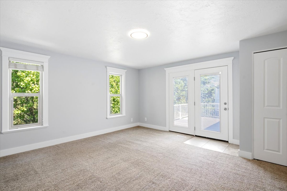 Neutral-toned carpeting extends throughout this room, complemented by two large windows with white trim and a double glass-panel door opening to a balcony with white railing