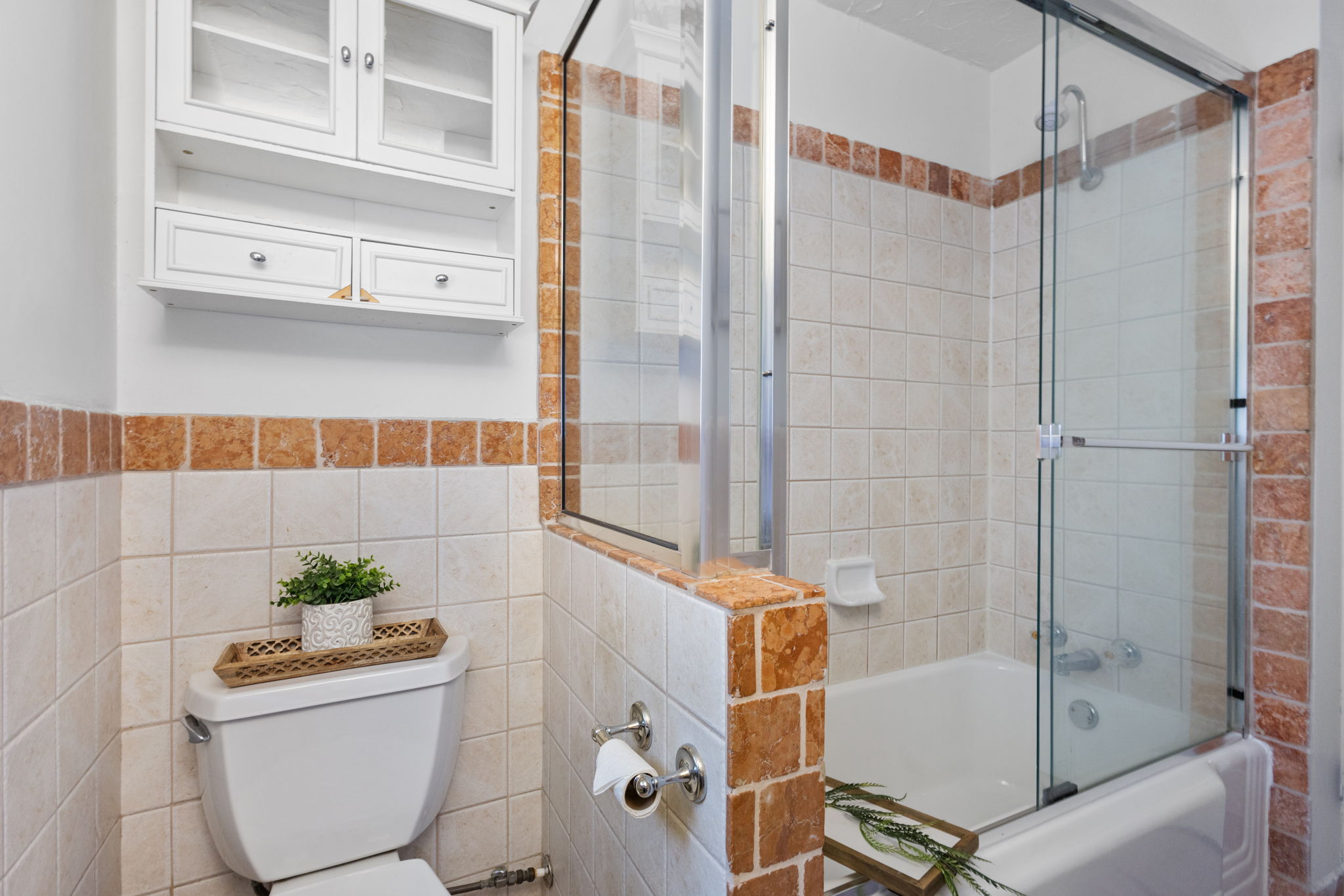 Bathroom featuring a built-in bathtub with glass sliding doors, white wall-mounted cabinetry with clear glass panels, and tiled walls with a decorative accent border