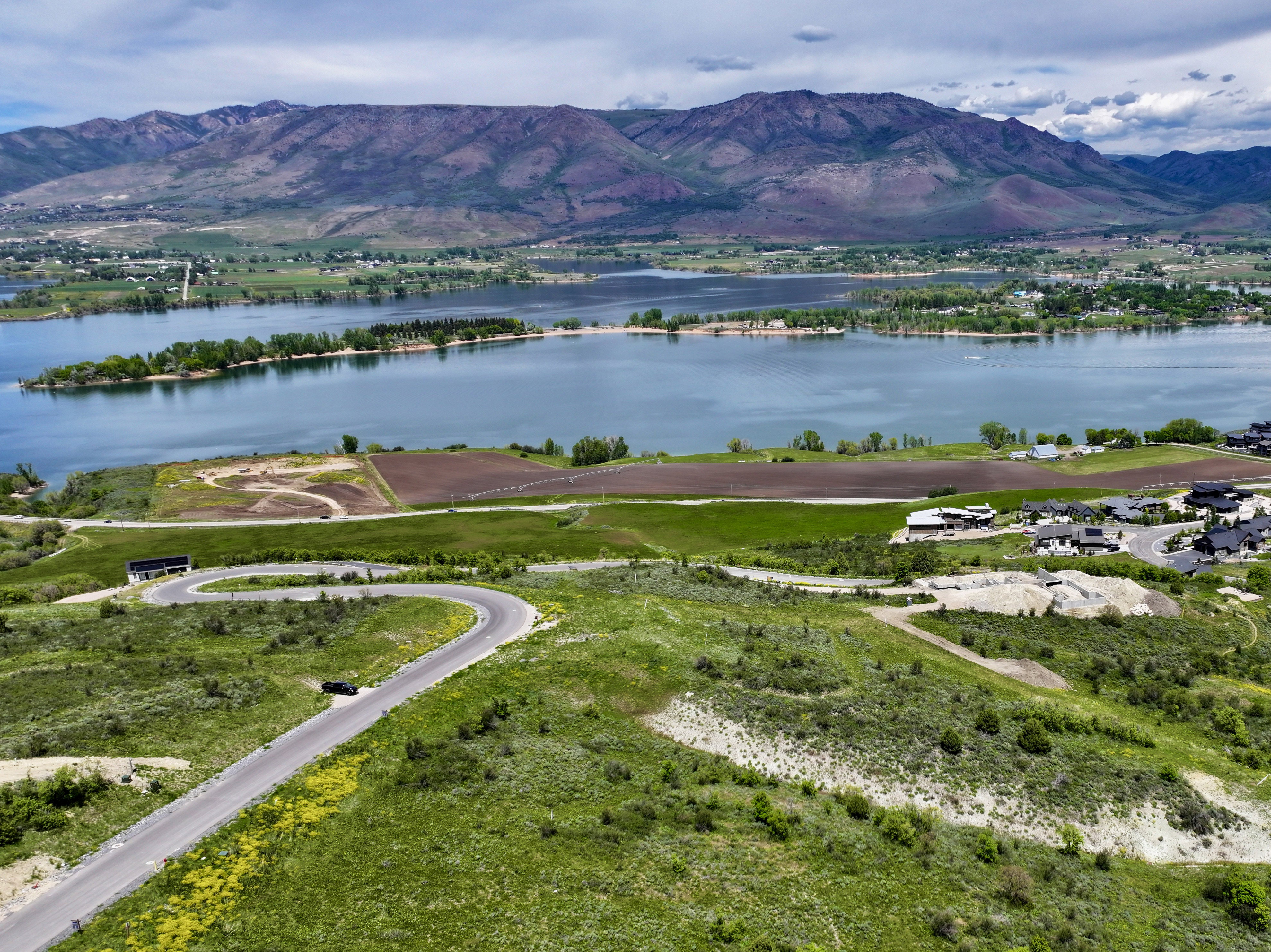 Expansive green landscape with a winding paved road, featuring a prominent lake backdrop with distant mountains, and a community of modern homes