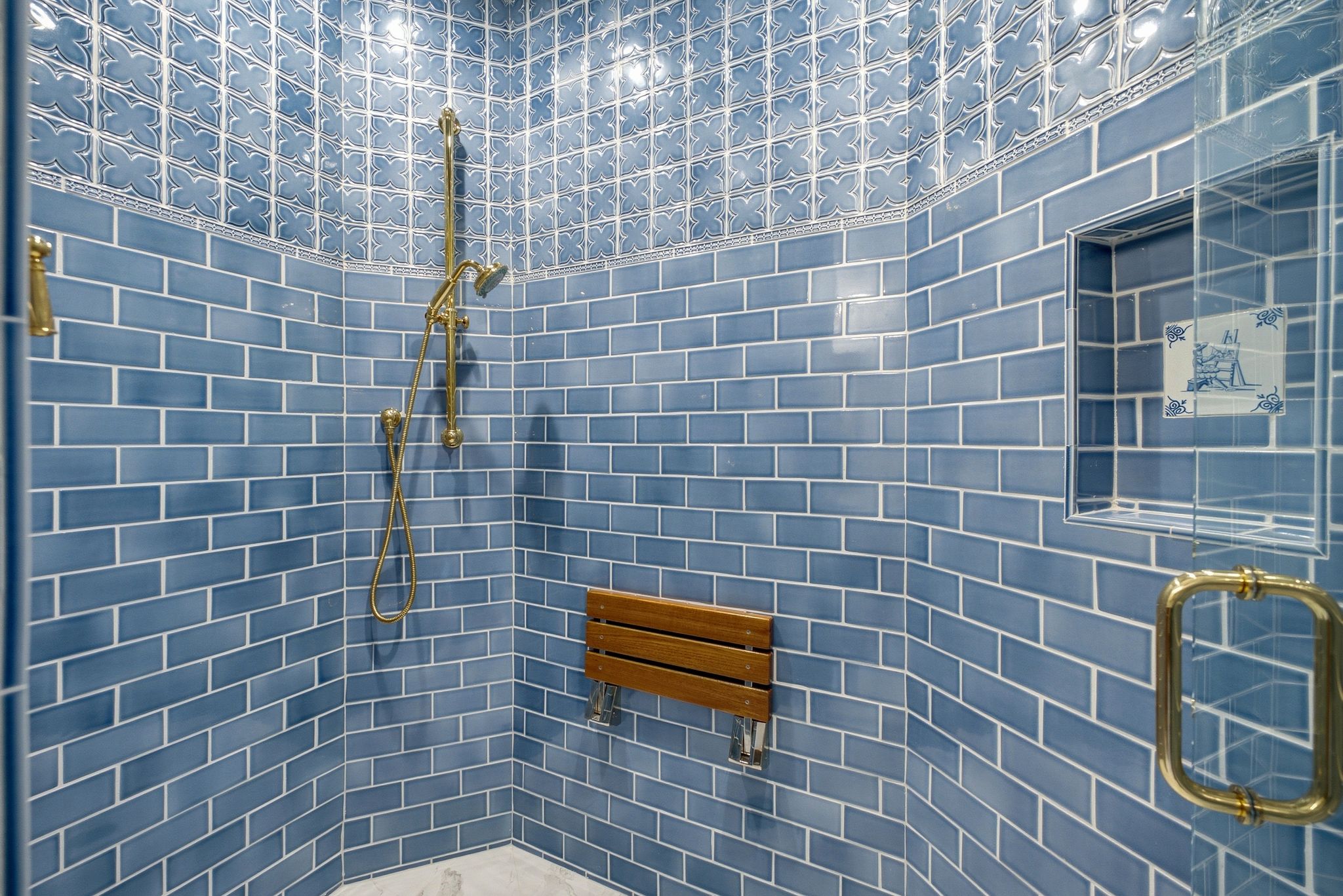 Shower with blue subway tile walls, patterned accent tile, brass fixtures, a built-in shower niche, and a wooden fold-down bench