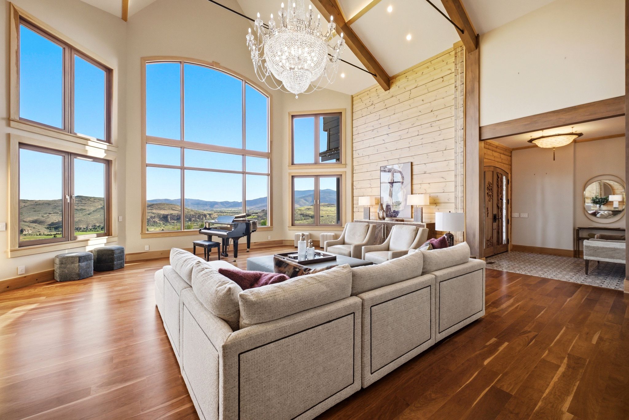 Two-story living area featuring extensive window walls, wood-finish flooring, exposed ceiling beams, a grand chandelier, and a wood-paneled accent wall