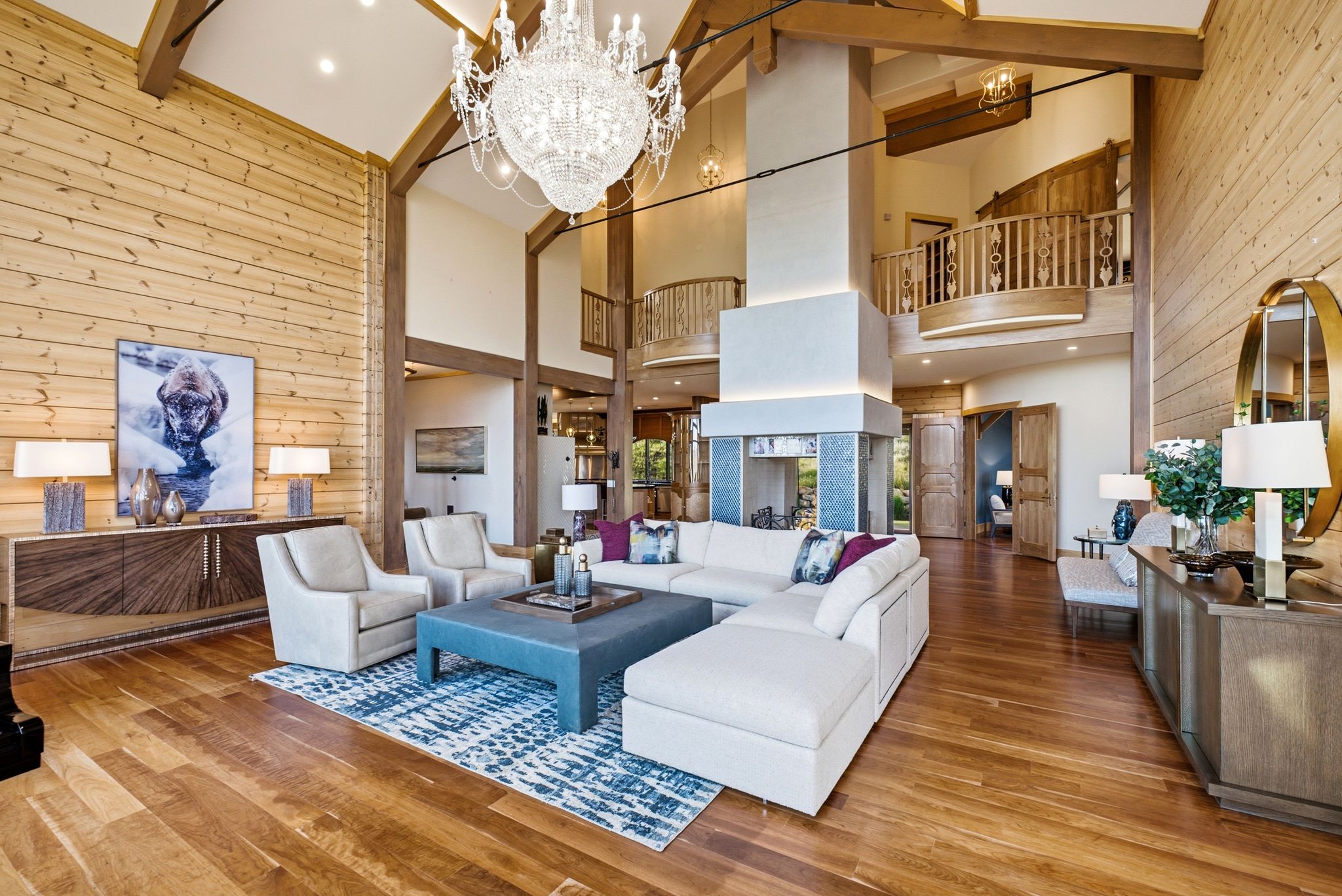 Vaulted ceiling living area featuring a prominent chandelier, exposed ceiling beams, wood-finish flooring, a central fireplace, and a second-story balcony