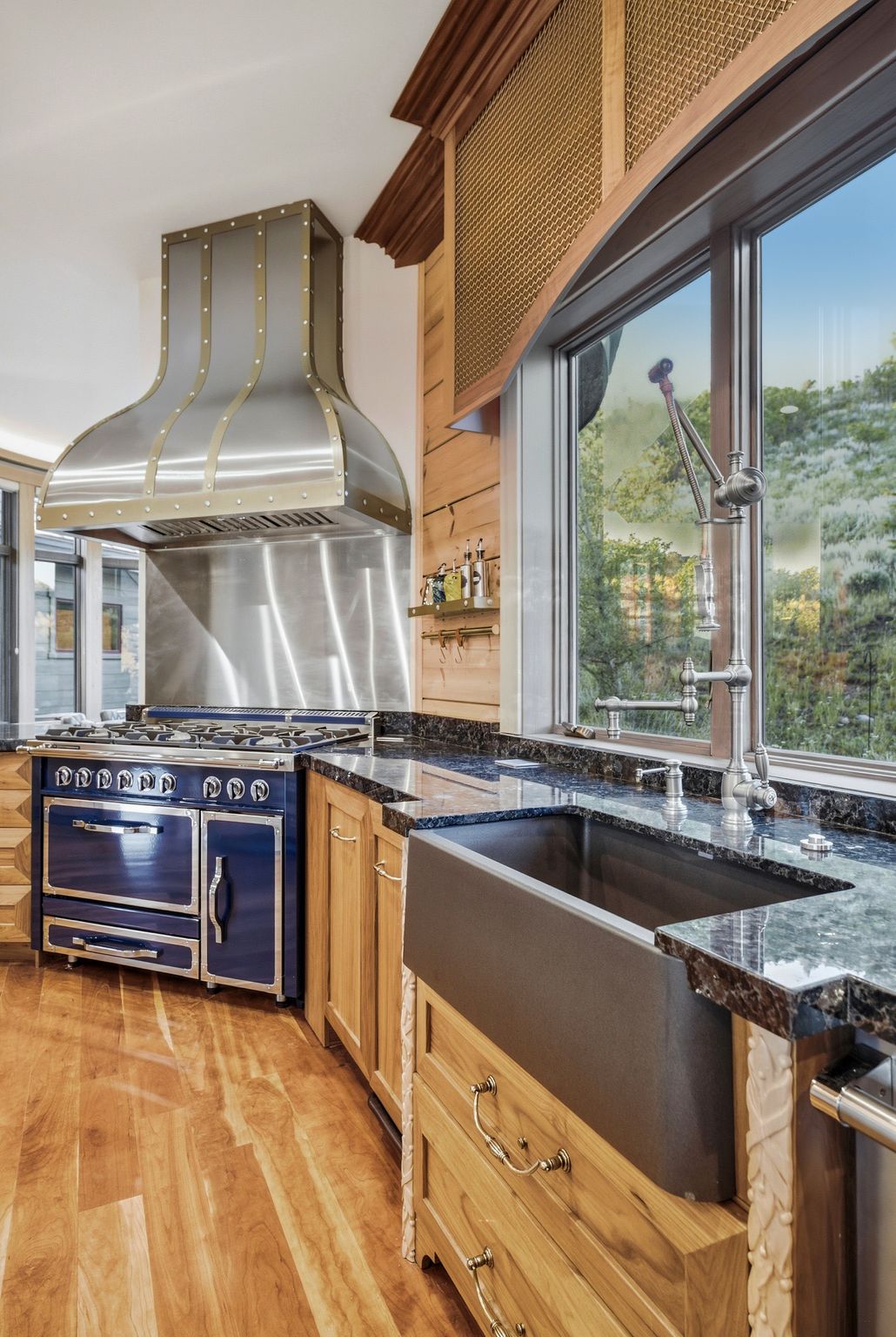 Professional-grade range with a custom stainless steel and brass hood, complemented by natural wood cabinetry, dark stone countertops, and a farmhouse-style sink with a gooseneck faucet