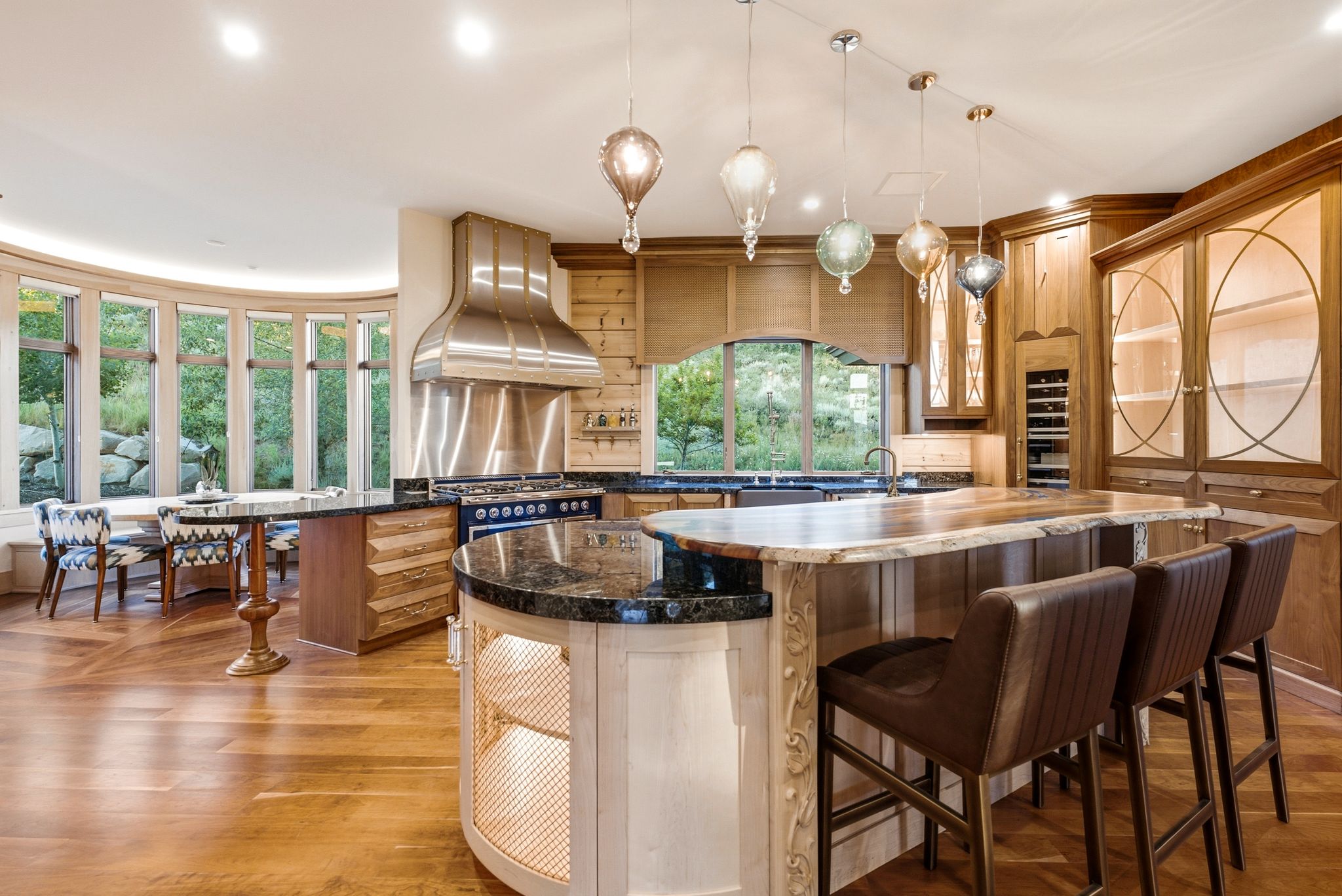 Gourmet kitchen featuring a large granite-topped island, wood-finish flooring, custom wood cabinetry, stainless steel range hood, and a curved window wall