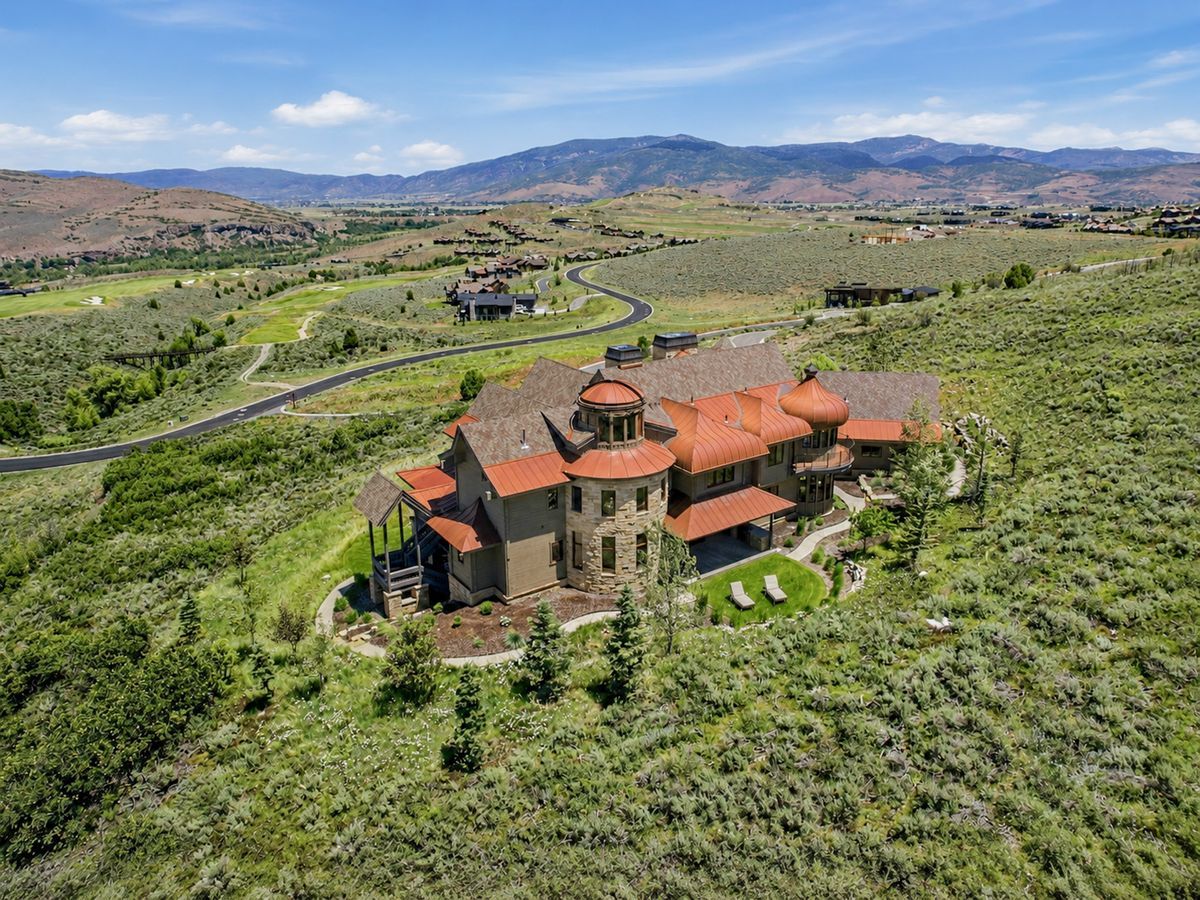 Stone and stucco exterior featuring a prominent circular turret with a domed copper roof, multiple additional copper-domed roof sections, a pitched shingle roof, and a stone retaining wall