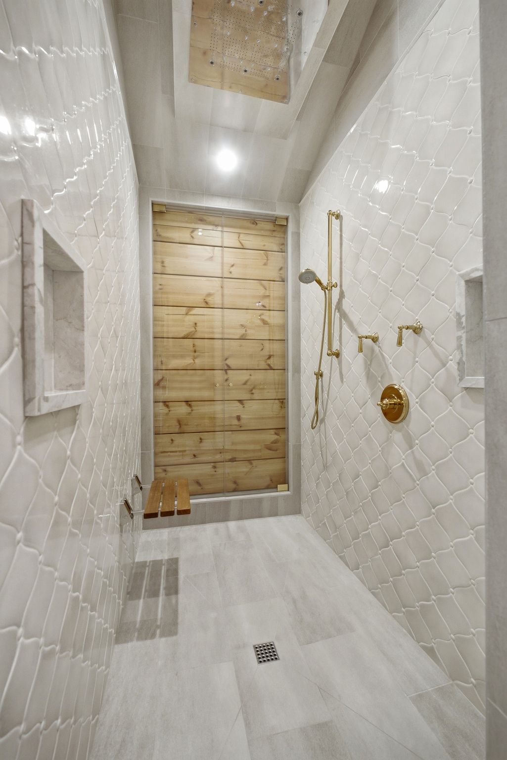 Spacious walk-in shower featuring light-toned tile walls with a textured pattern, a built-in wooden bench, a recessed wall niche, and warm-toned metallic shower fixtures
